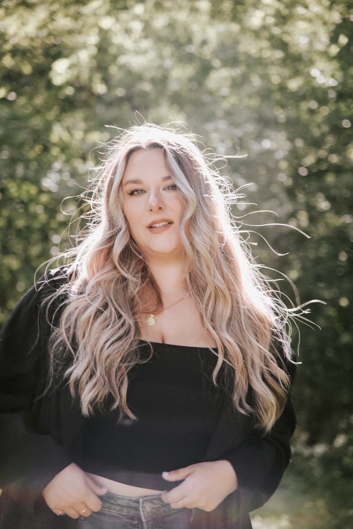 Young woman with long, wavy blonde hair outdoors, sunlight filtering through trees, wearing a black top and jacket.