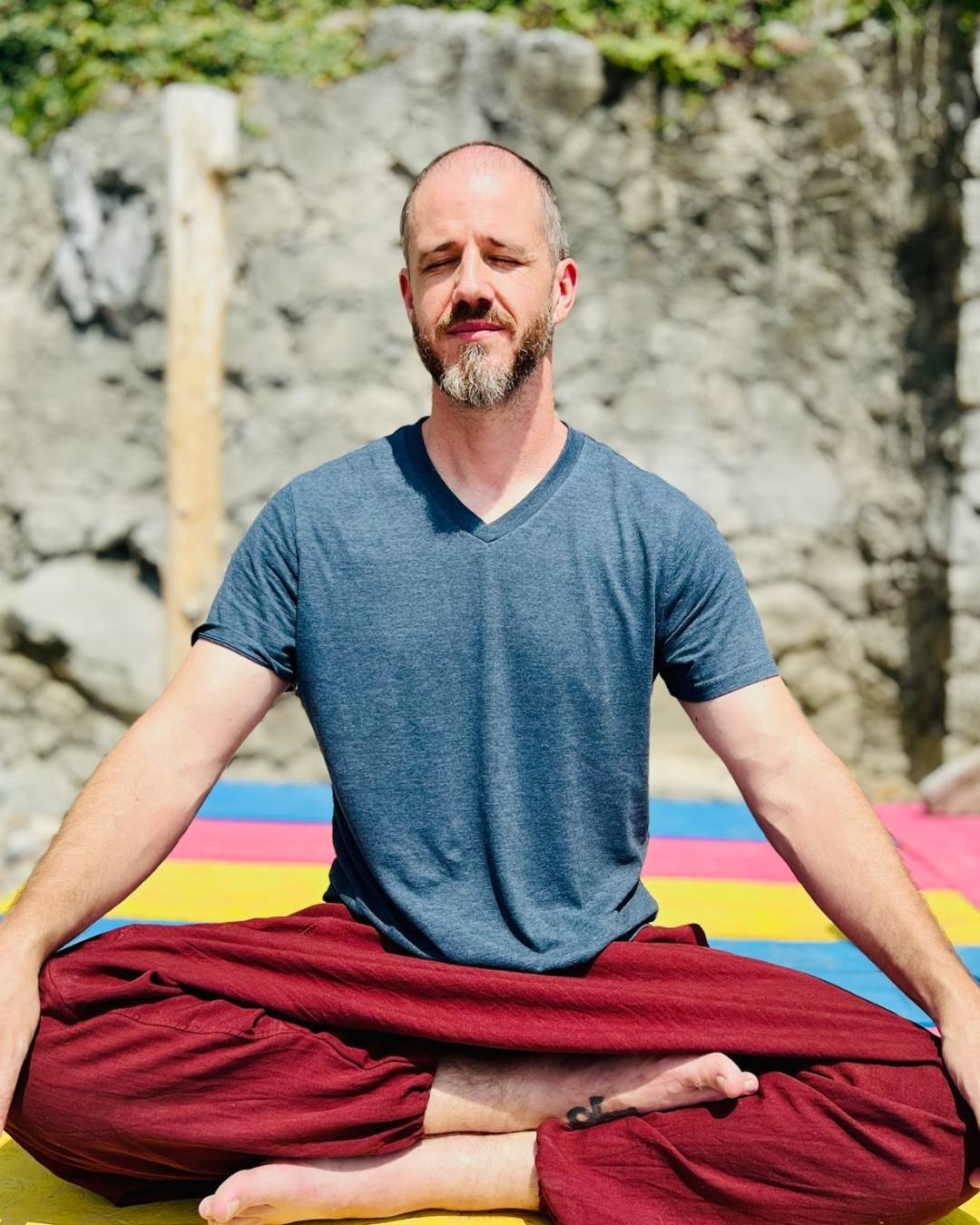Man sitting cross-legged outdoors with colorful mat, meditating with eyes closed, stone wall and greenery behind him.