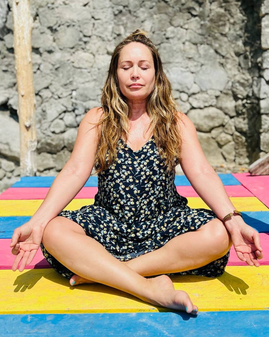 Woman sitting cross-legged on colorful mats, meditating outdoors against a stone wall, eyes closed, peaceful expression.