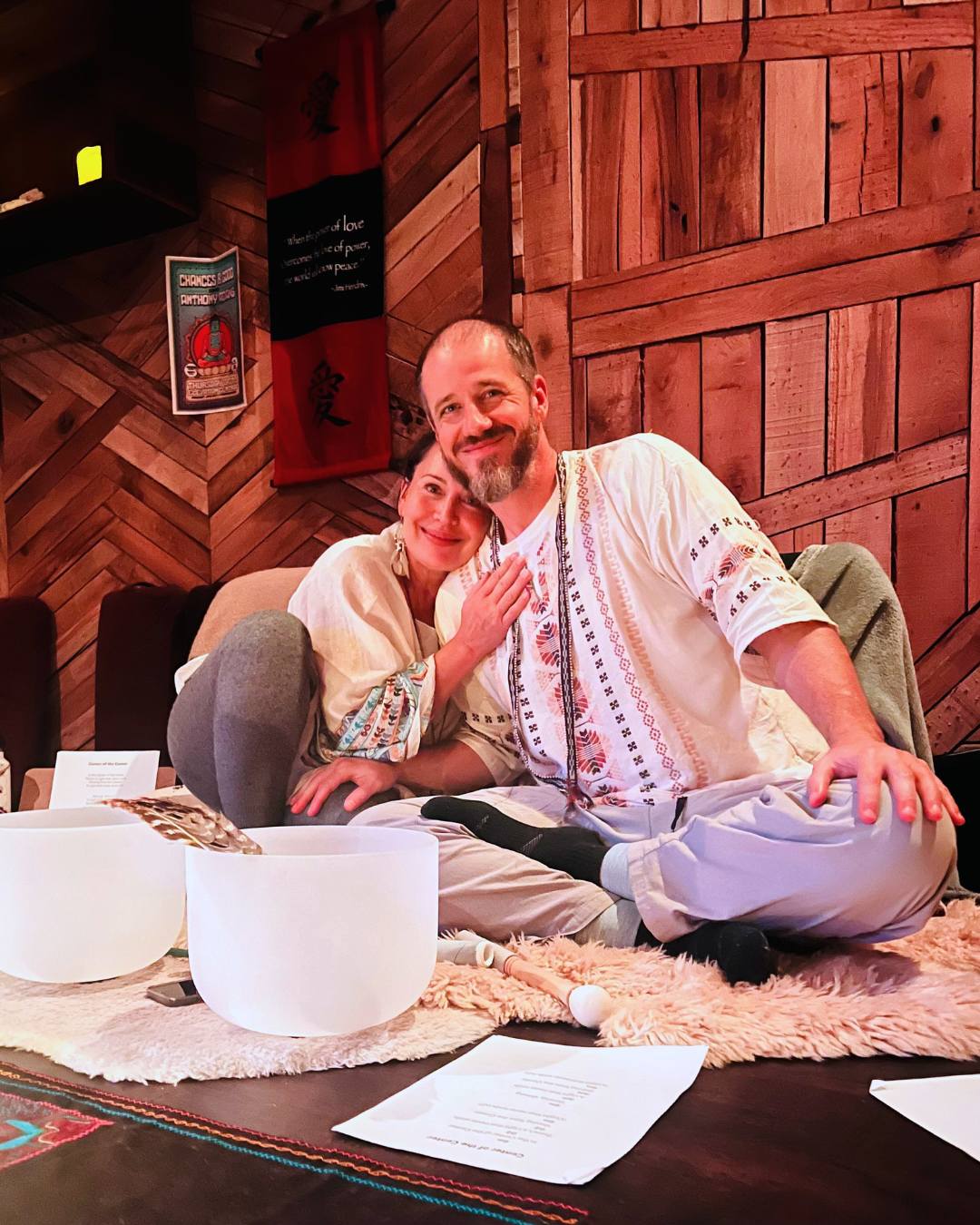 Two people sitting on a rug, smiling, with wooden wall background, musical instruments, and papers in front.