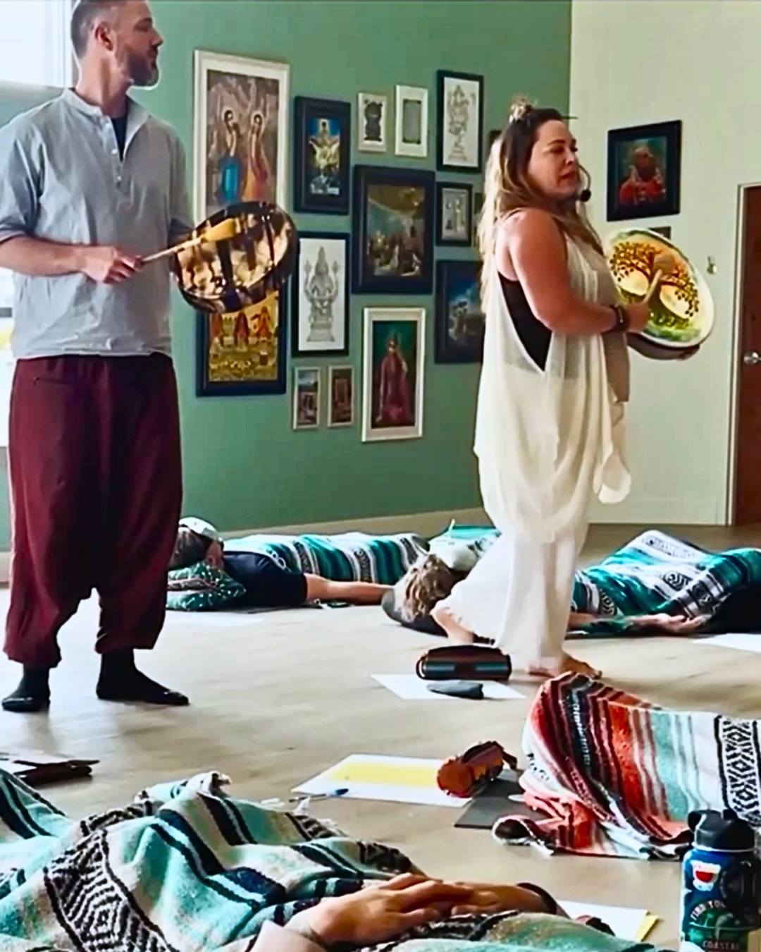 Two people in a room with art on the wall, one holding a drum, the other with a plate, cushions on the floor.
