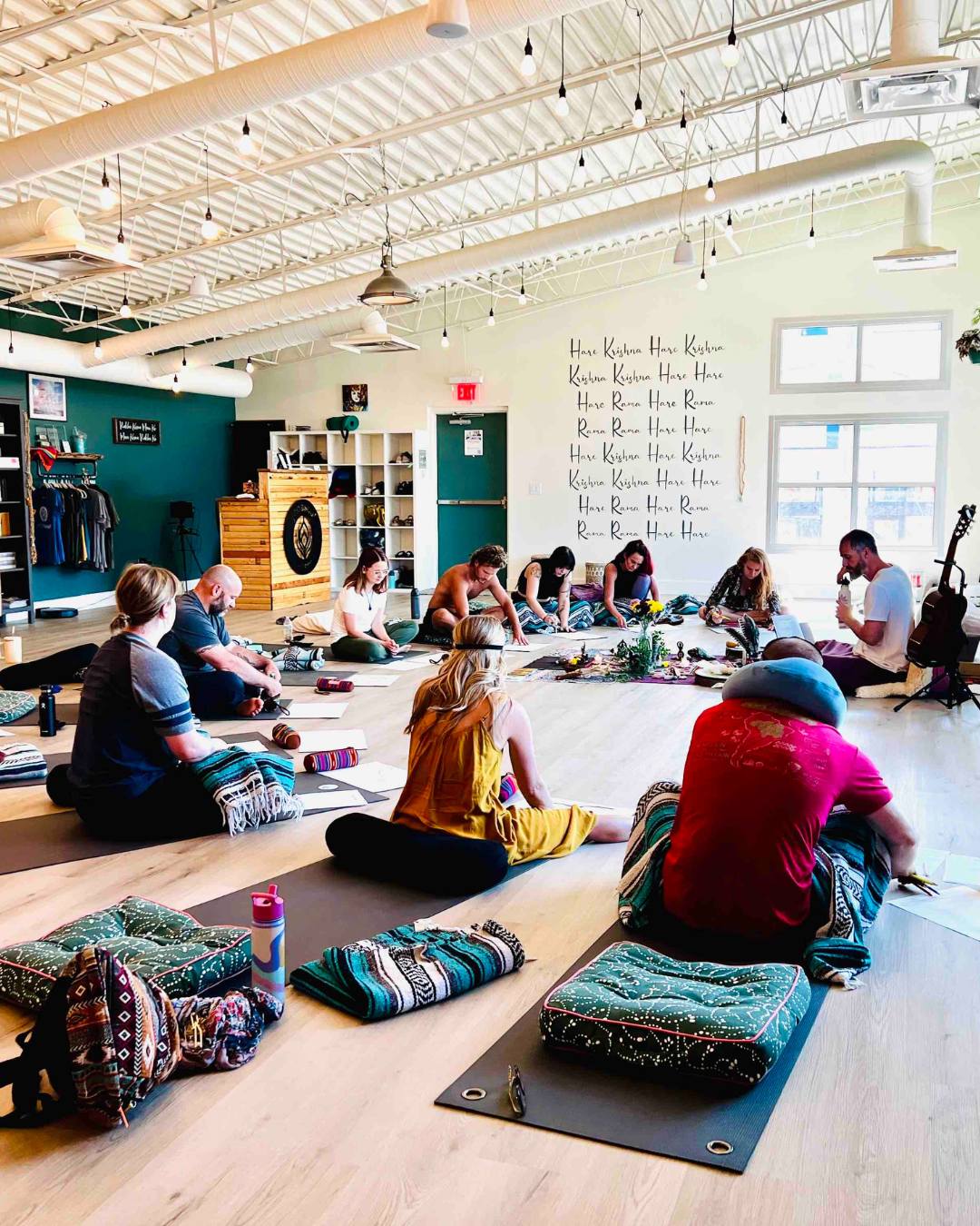 People sitting on mats in a spacious room with high ceiling, large windows, and a wall with text and decorations.