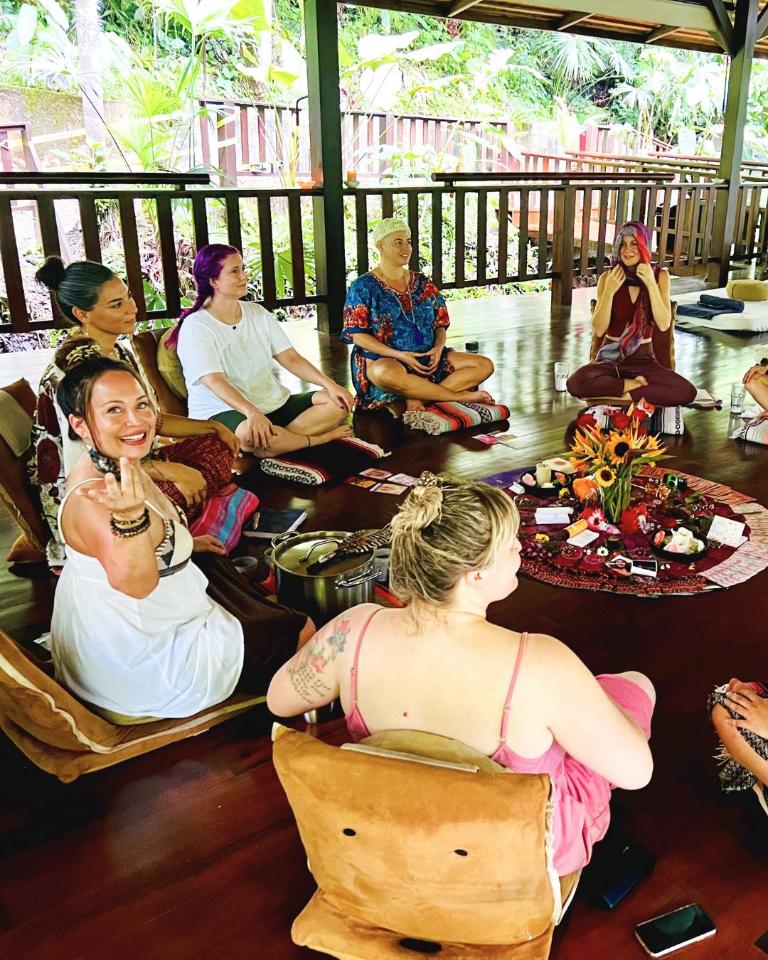 Group of people sitting on cushions and chairs in a covered outdoor area, with a table of flowers and items in front.