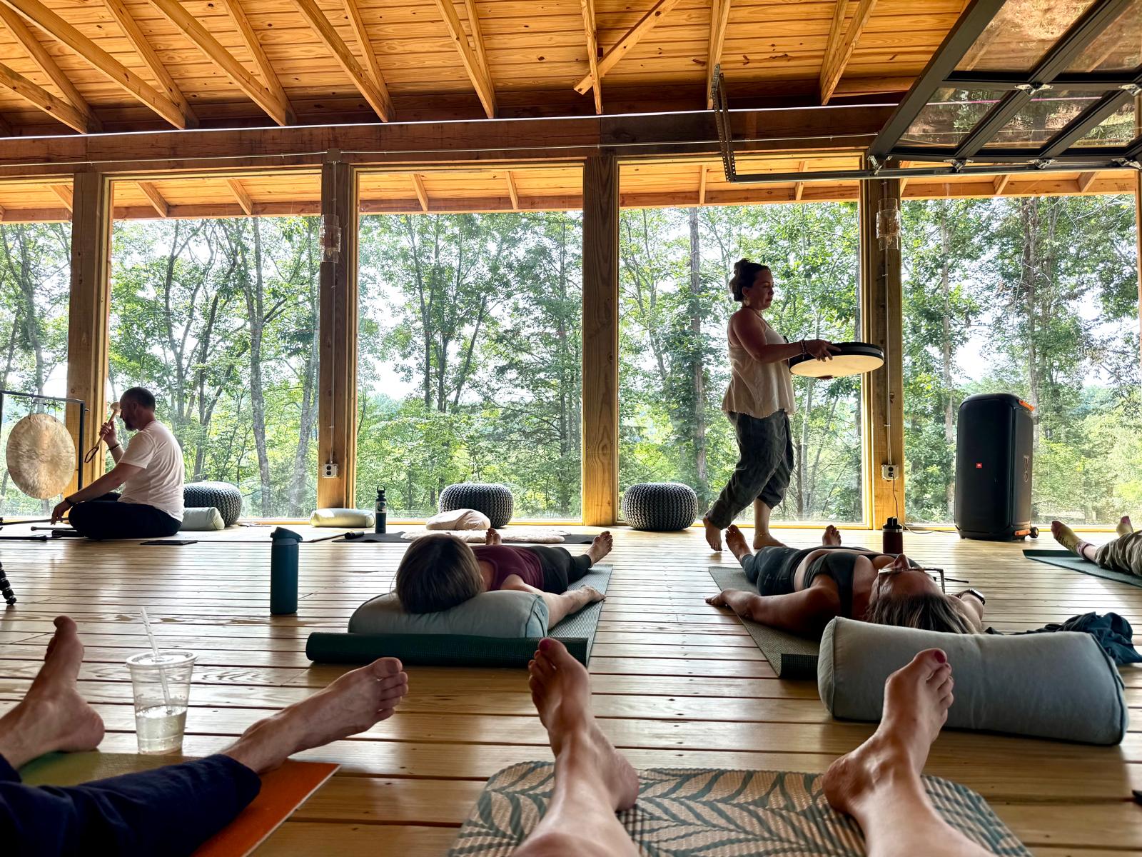 People practicing yoga on mats in a bright, open studio with large windows and trees outside.