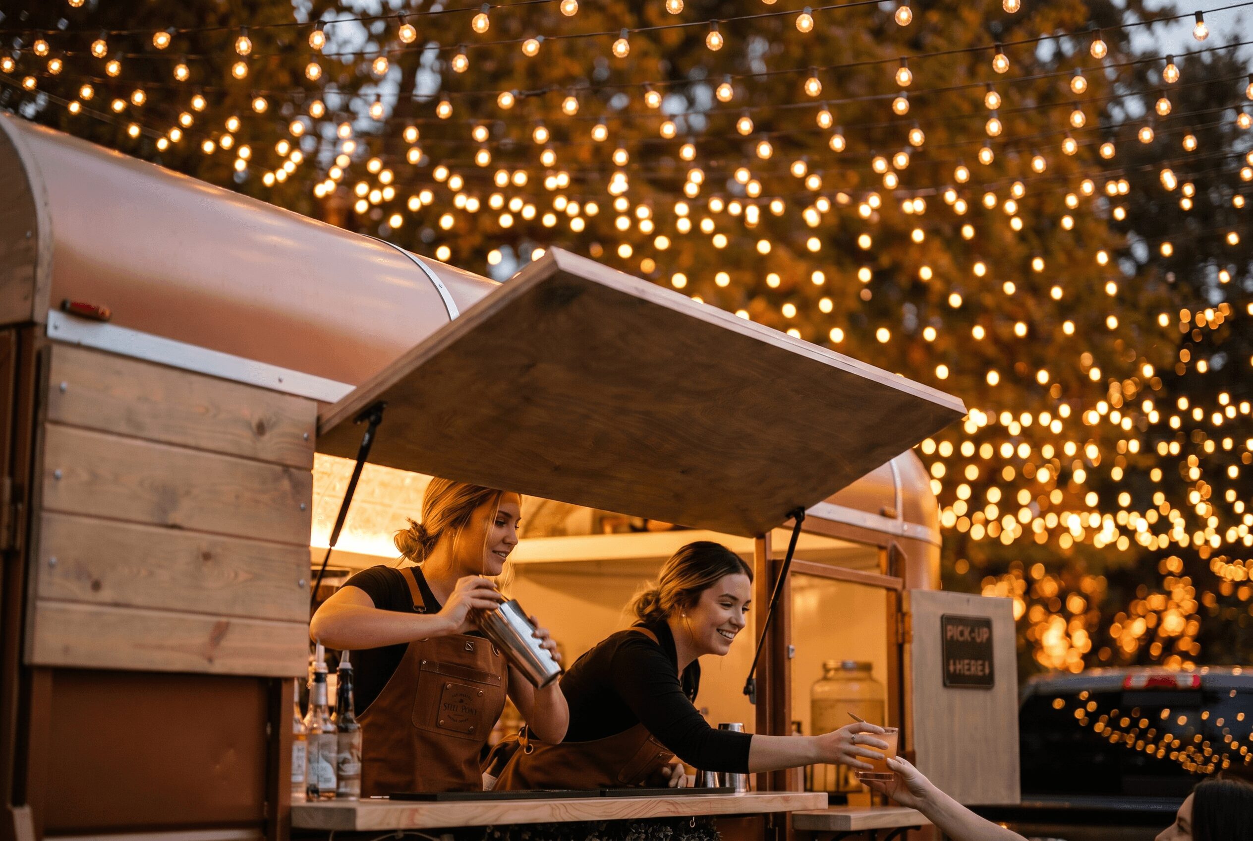 Two women working at a food stand with string lights in the background, one pouring a drink and the other serving.