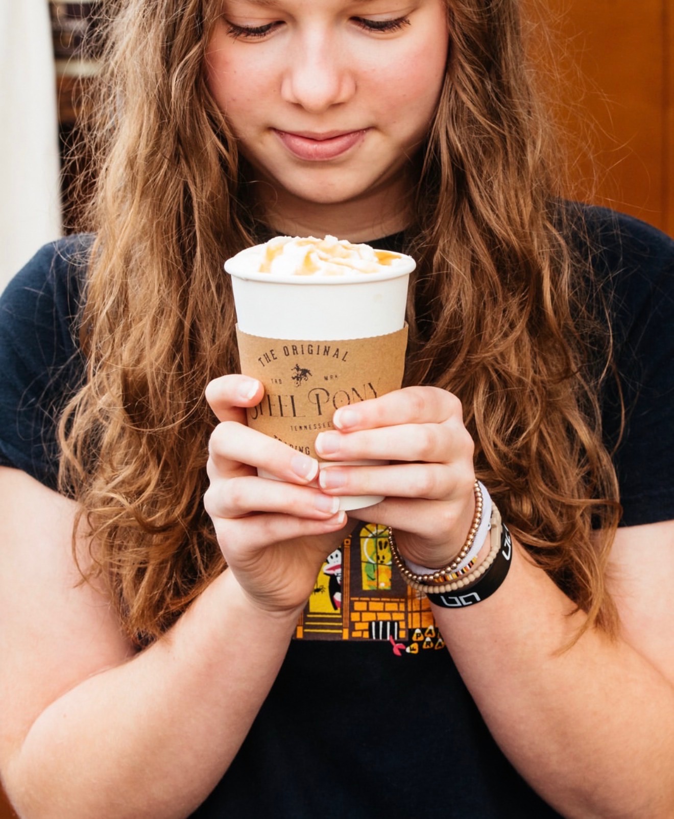Girl holding a cup of ice cream with both hands, looking at it, wearing a black shirt and bracelets.