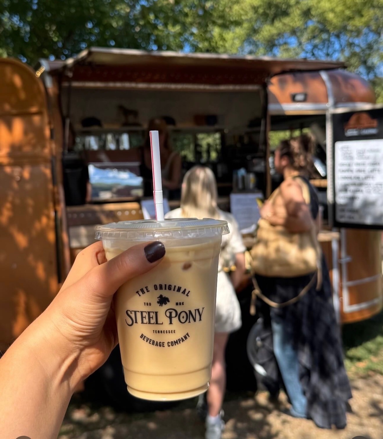 Hand holding a plastic cup with a straw, in front of a food truck with people outside, trees in background.