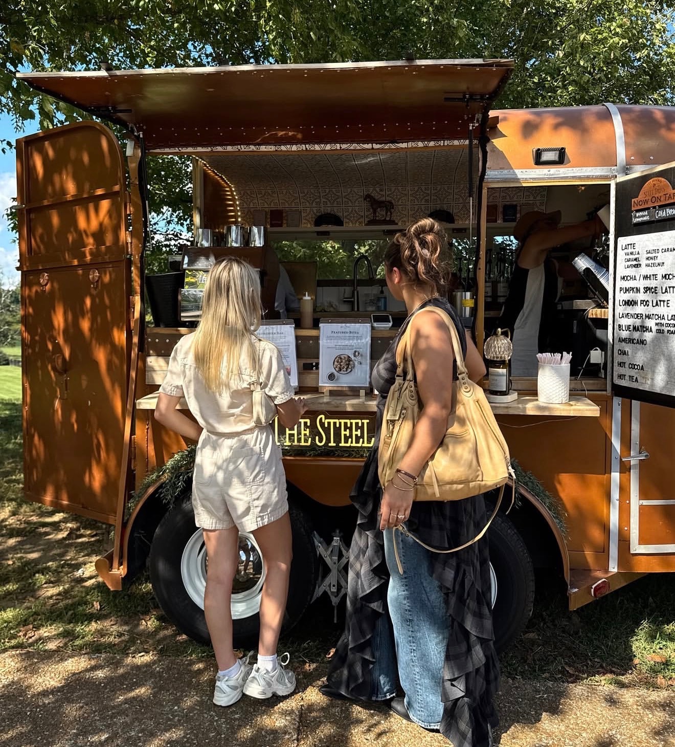 Two women stand at a food truck outdoors, one with long blonde hair and the other with dark hair, both facing the truck.