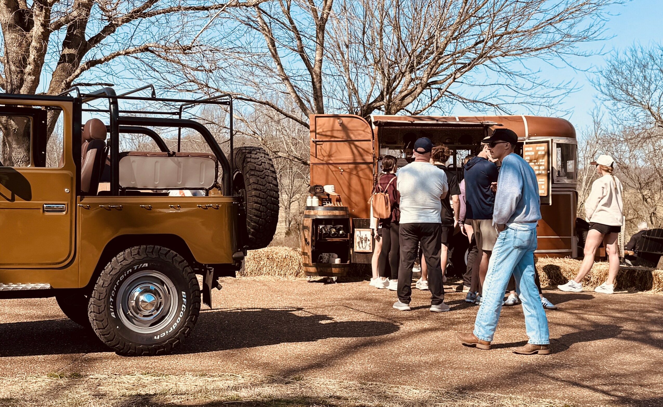 Group of people standing near vintage vehicles outdoors on a sunny day, with leafless trees in the background.