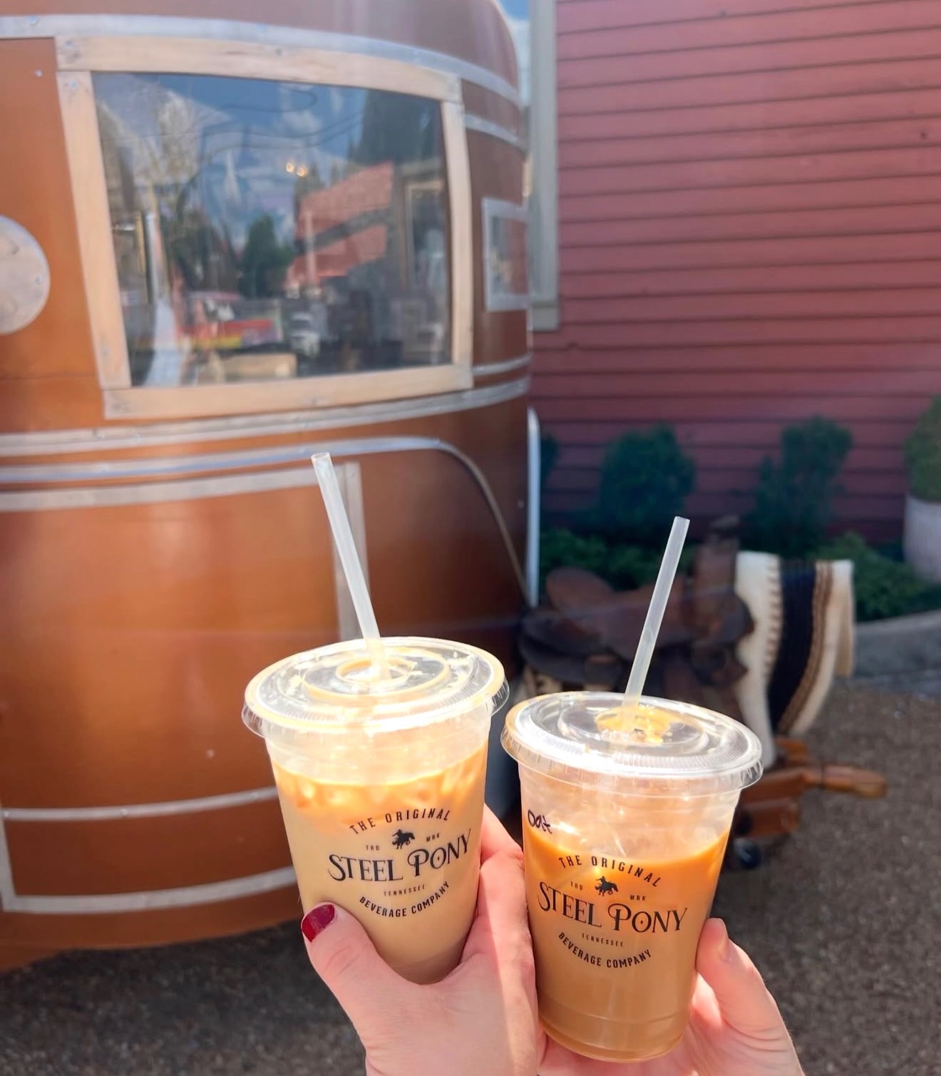 Two iced drinks in plastic cups with straws held up in front of a vintage trailer and pink wall, outdoor setting.