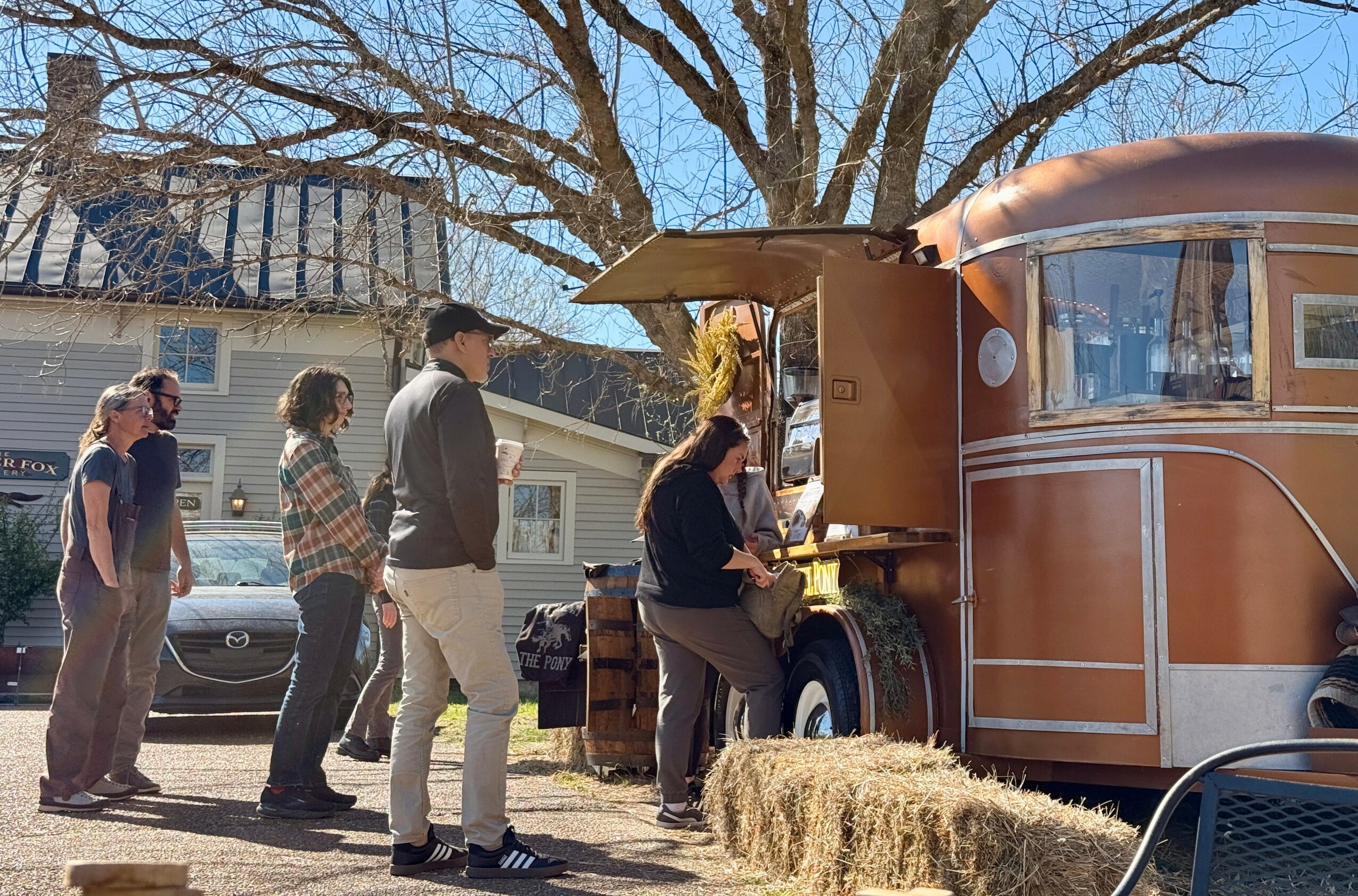 People line up at a food truck outdoors on a sunny day, with a tree and house in the background.