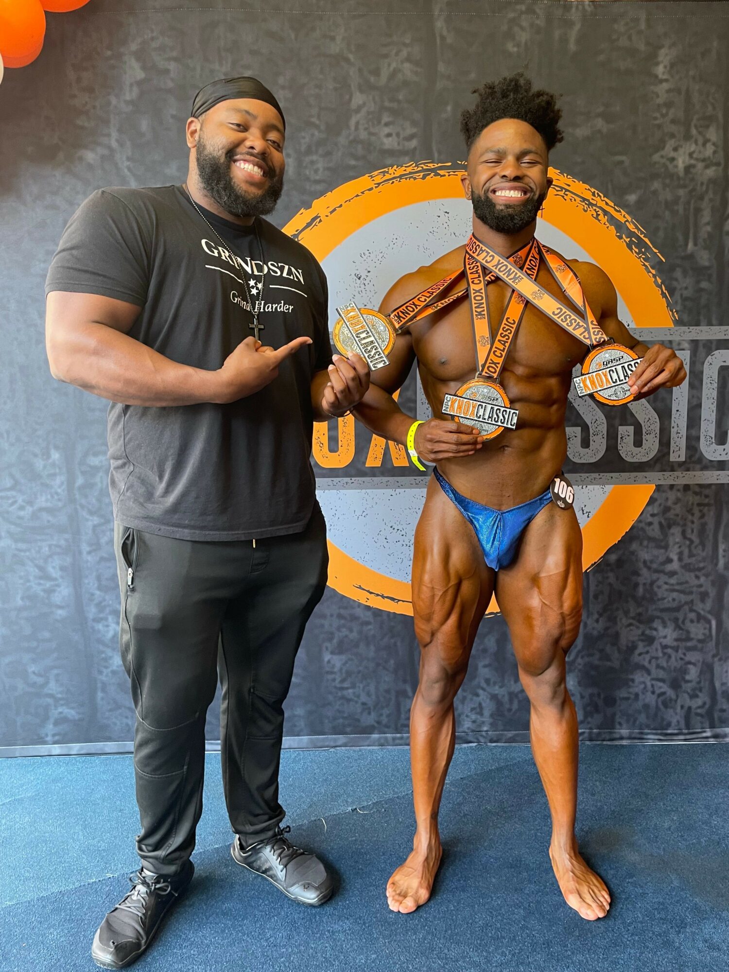 A muscular man in black pants and a black t-shirt stands next to a female athlete holding medals, in a gym setting.