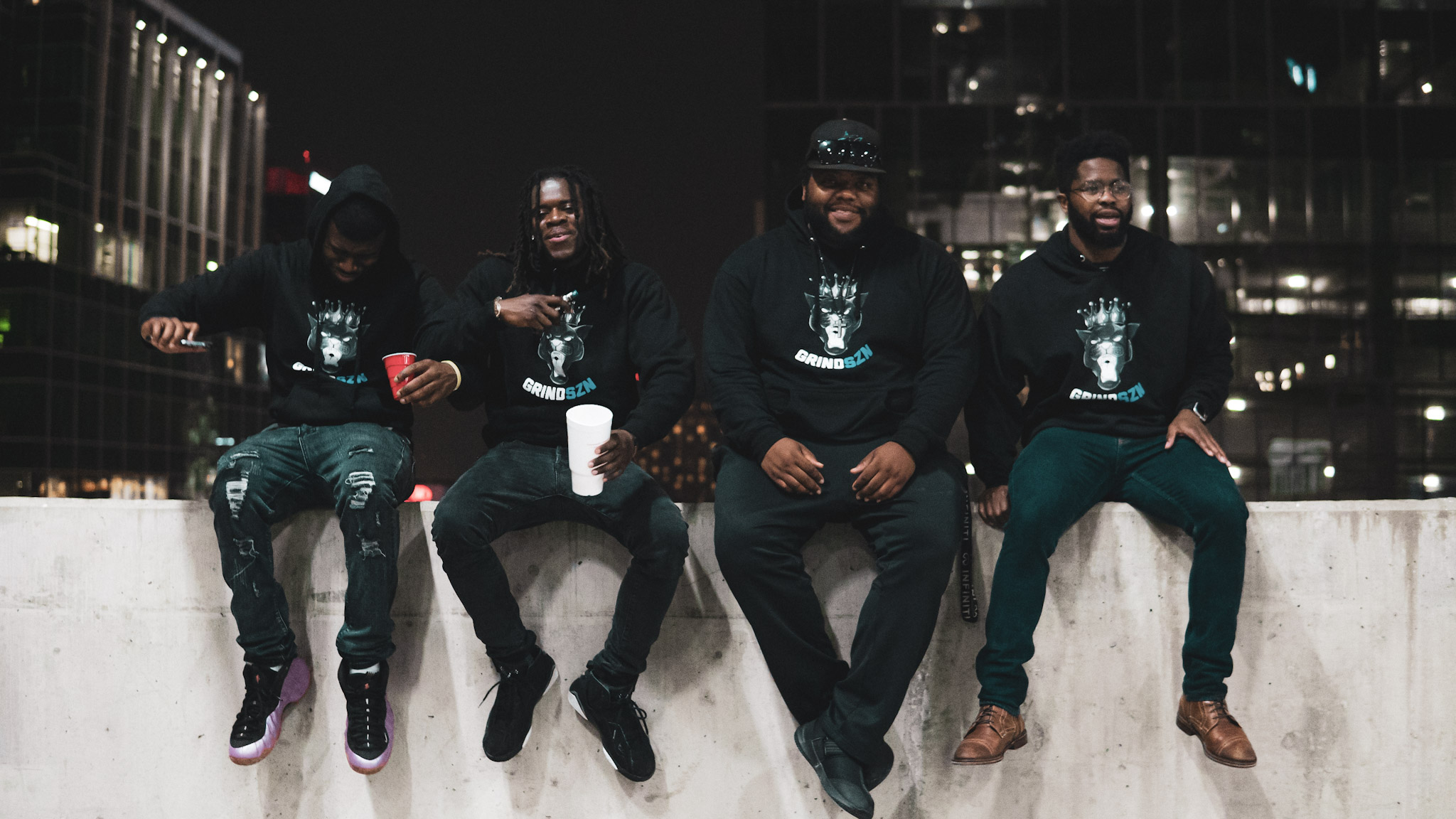 Four people sitting on a ledge at night with city buildings in the background, holding drinks and smiling.
