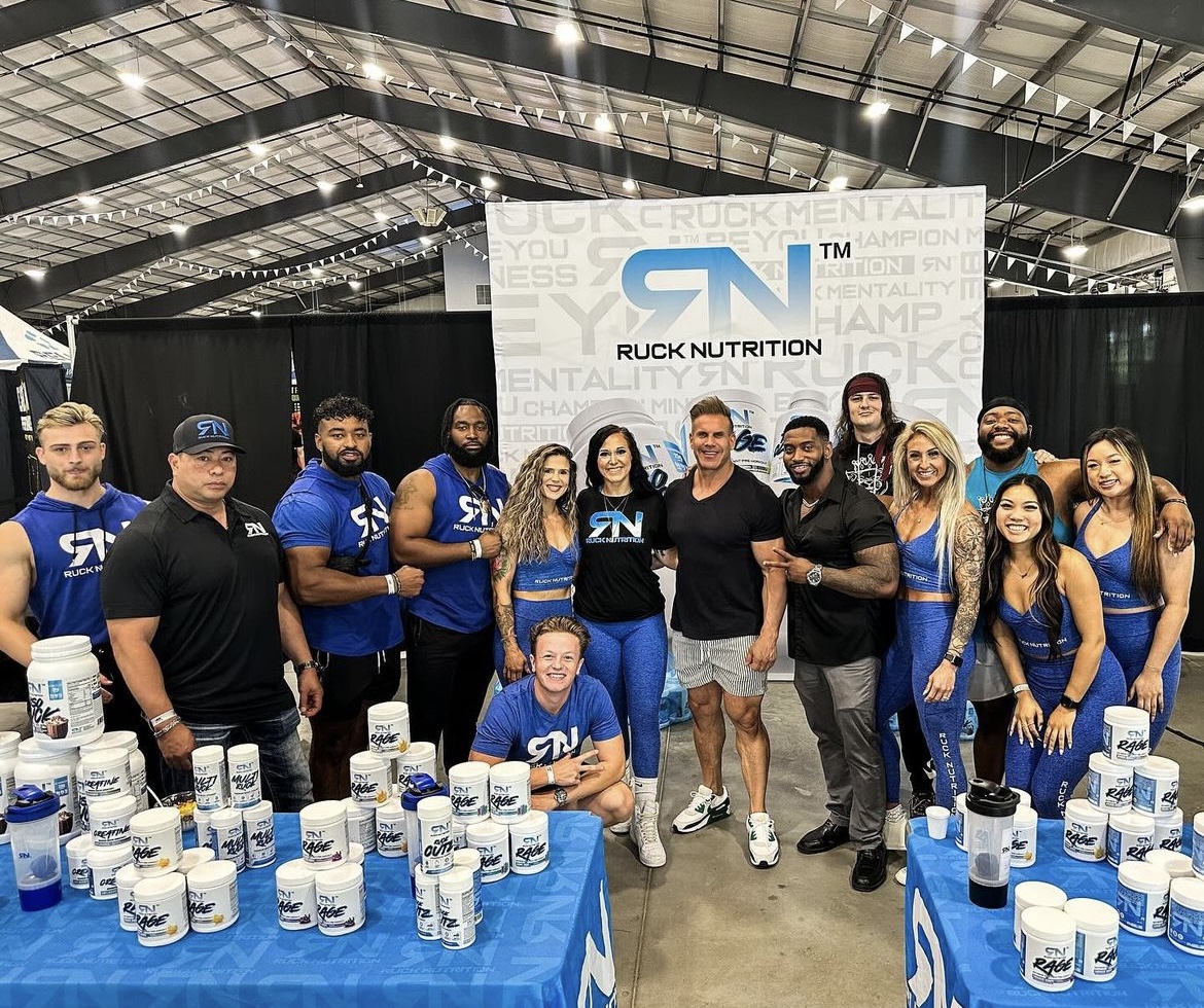 Group of people at a nutrition booth with containers and a large banner, inside a spacious indoor venue.