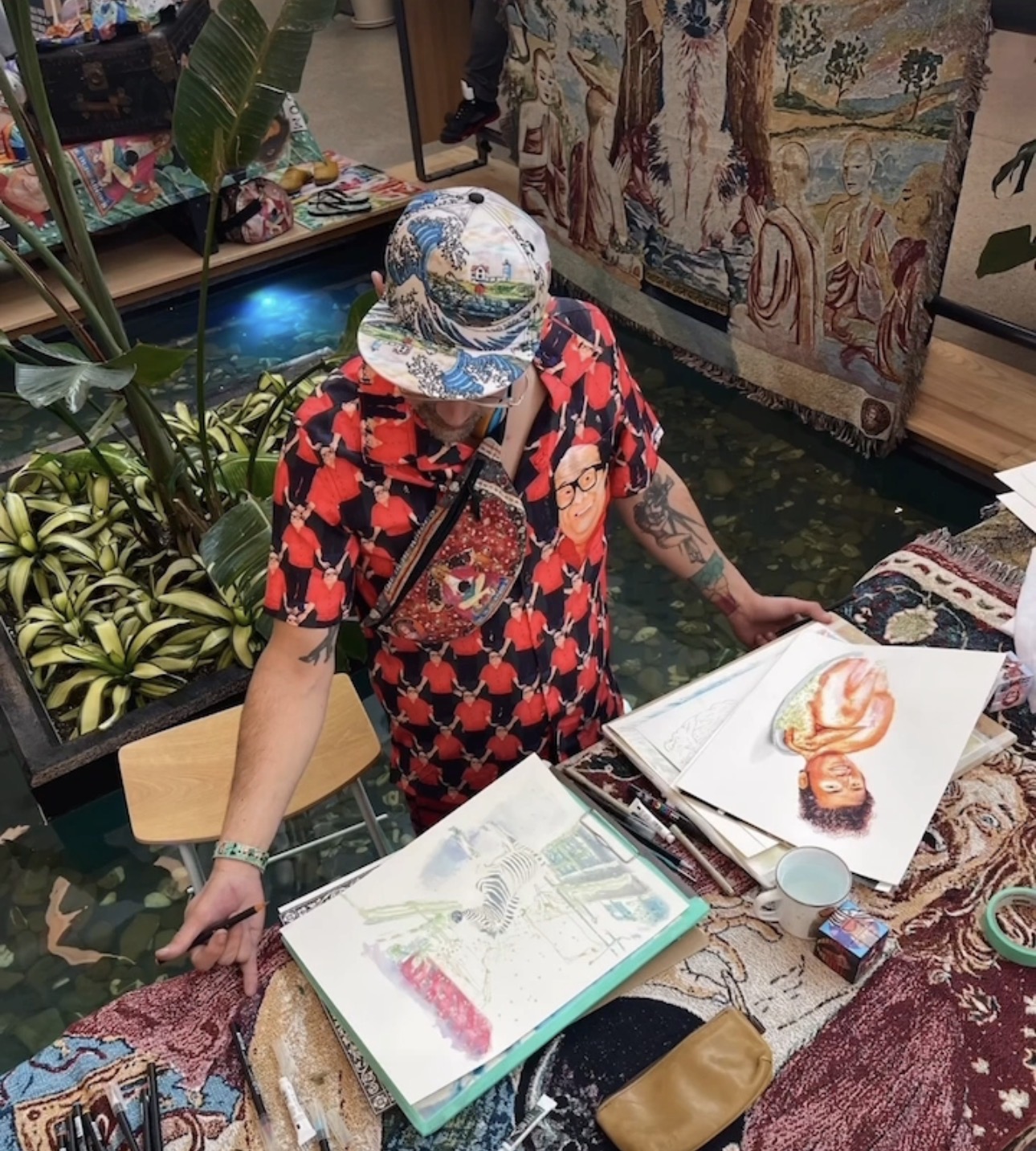 Person wearing a colorful hat and red patterned shirt looking at open sketchbooks on a table, surrounded by plants and artwork.