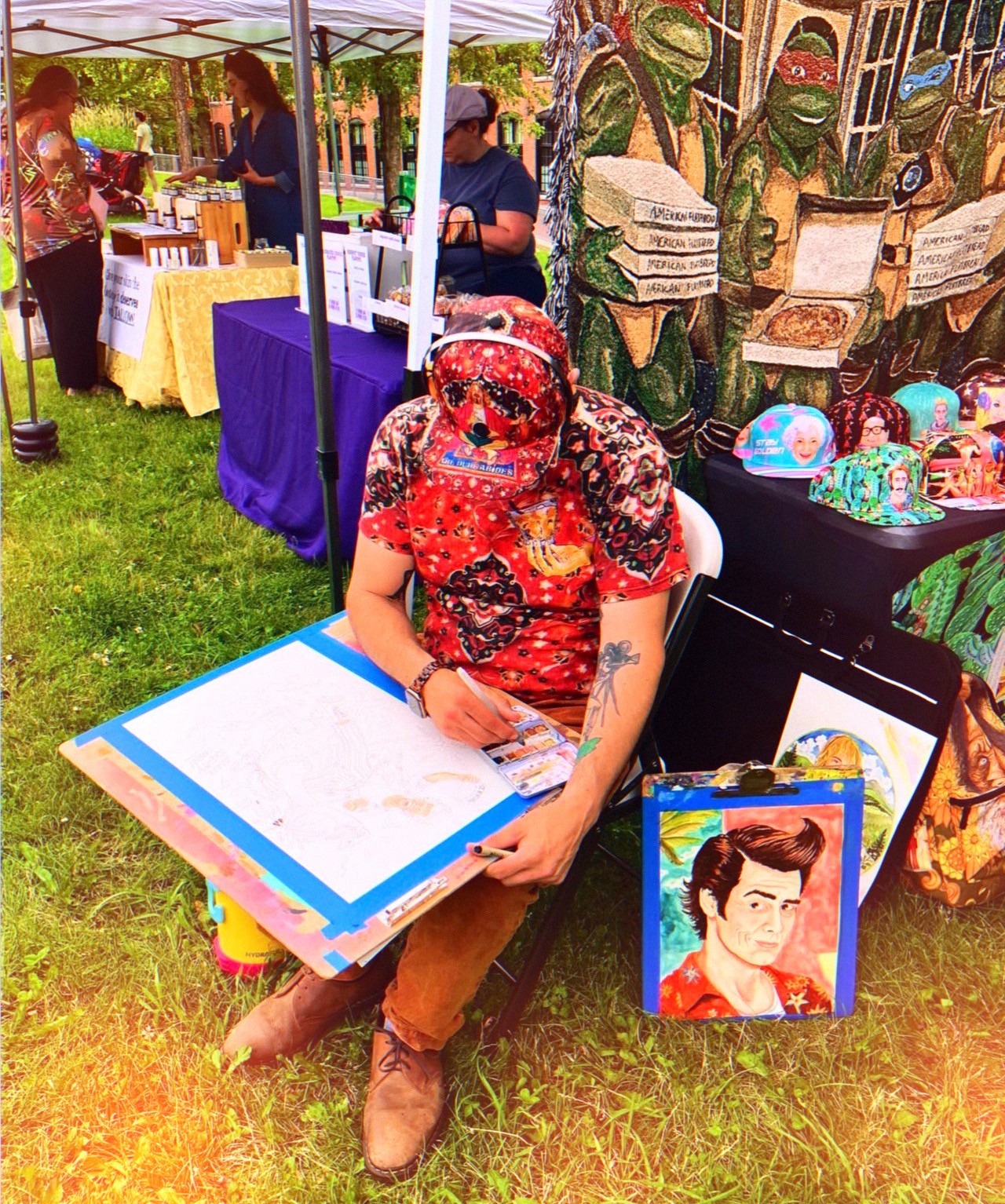 Person with a red floral mask sitting at a table with artwork and colorful items, outdoors at a market or fair.