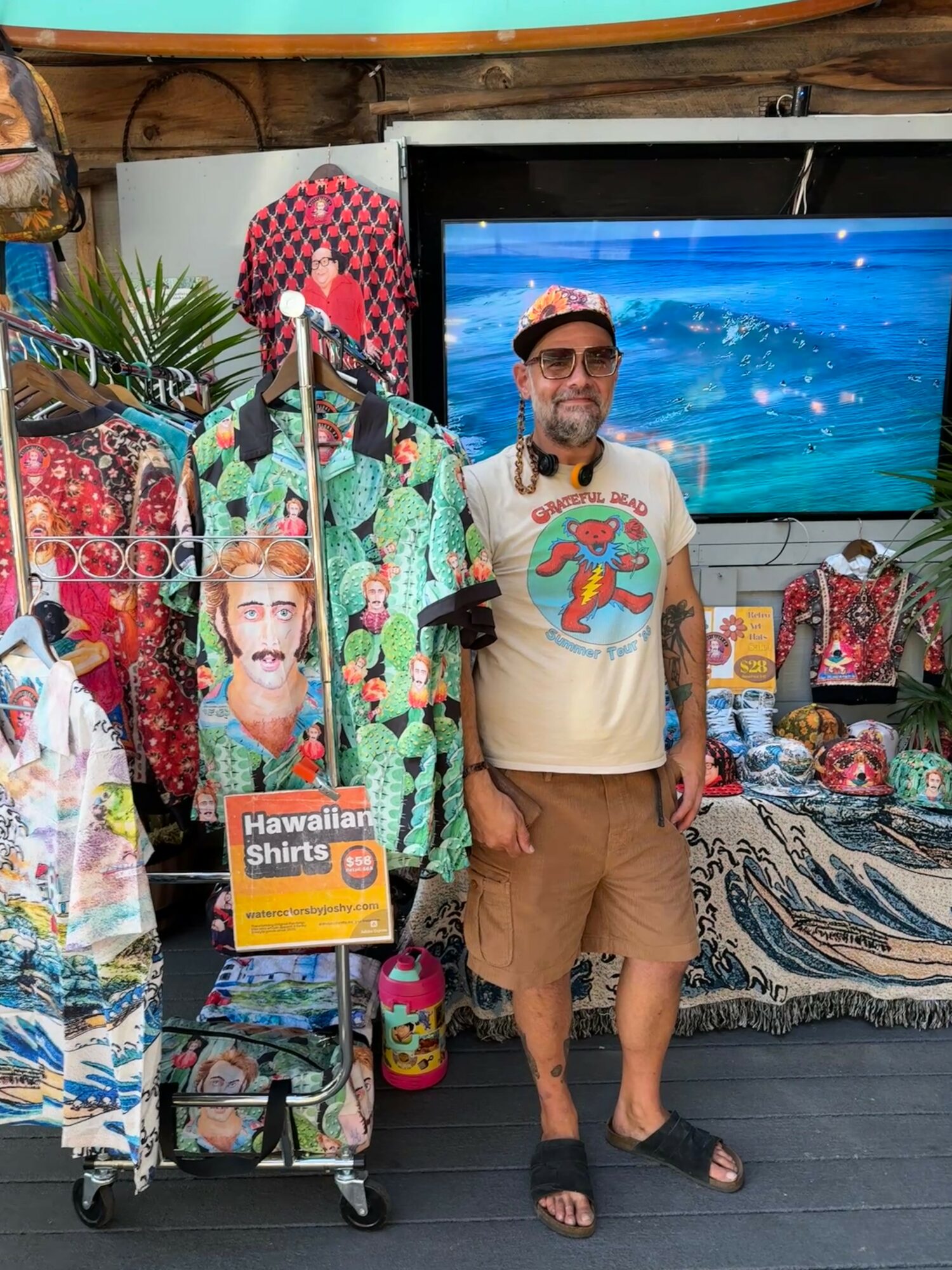 Man wearing sunglasses, cap, and Hawaiian shirt stands in front of Hawaiian shirts and a TV display, with a table of souvenirs nearby.