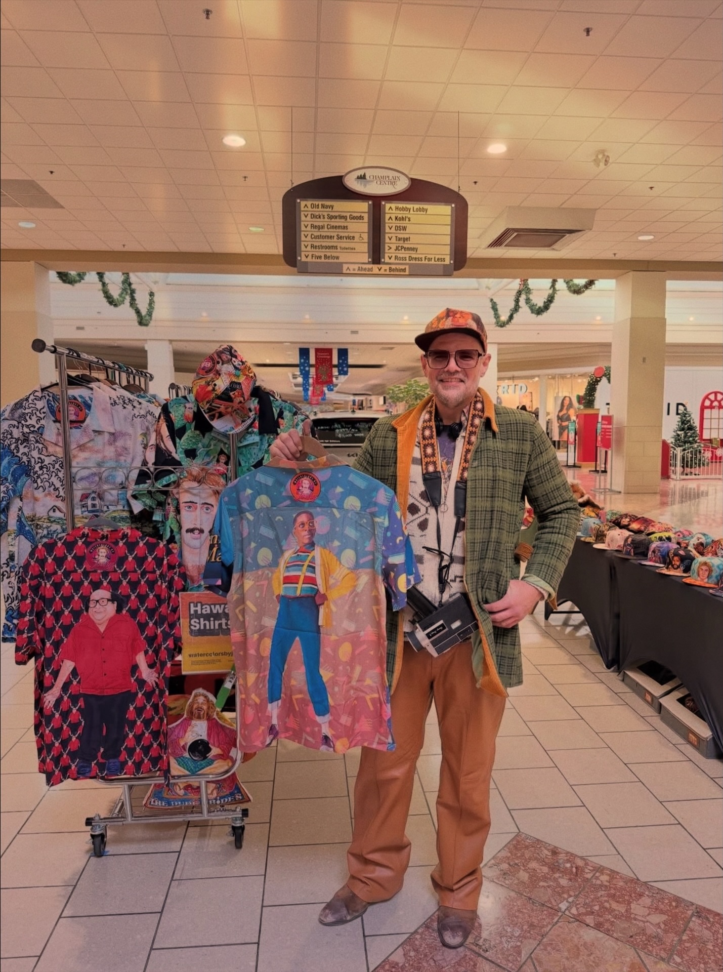 Person wearing sunglasses and a hat stands next to colorful t-shirts at a market stall, with a menu board overhead.