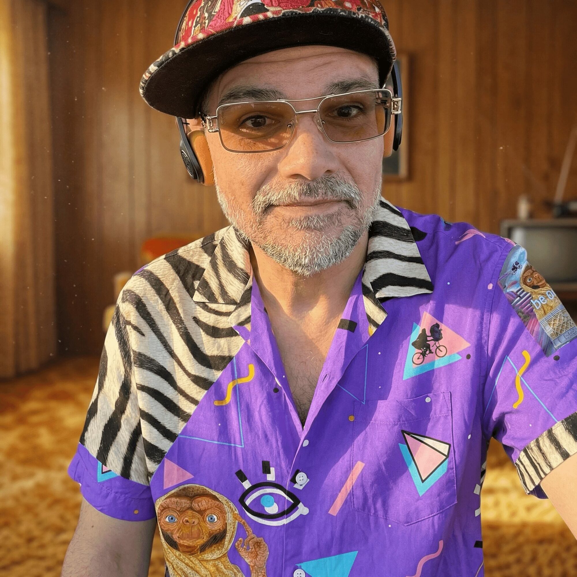 Man wearing glasses, a colorful hat, and a vibrant shirt with various patterns and illustrations, indoors with wooden walls.