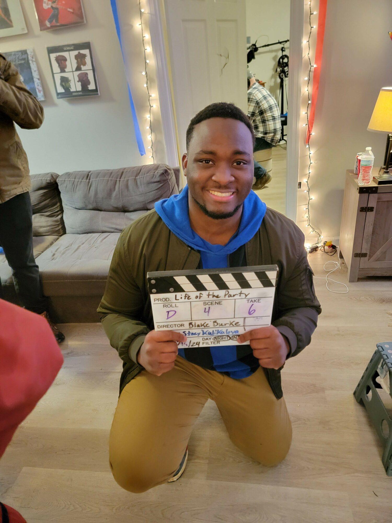 Person sitting on the floor holding a clapperboard, smiling, in a decorated room with string lights and framed pictures.