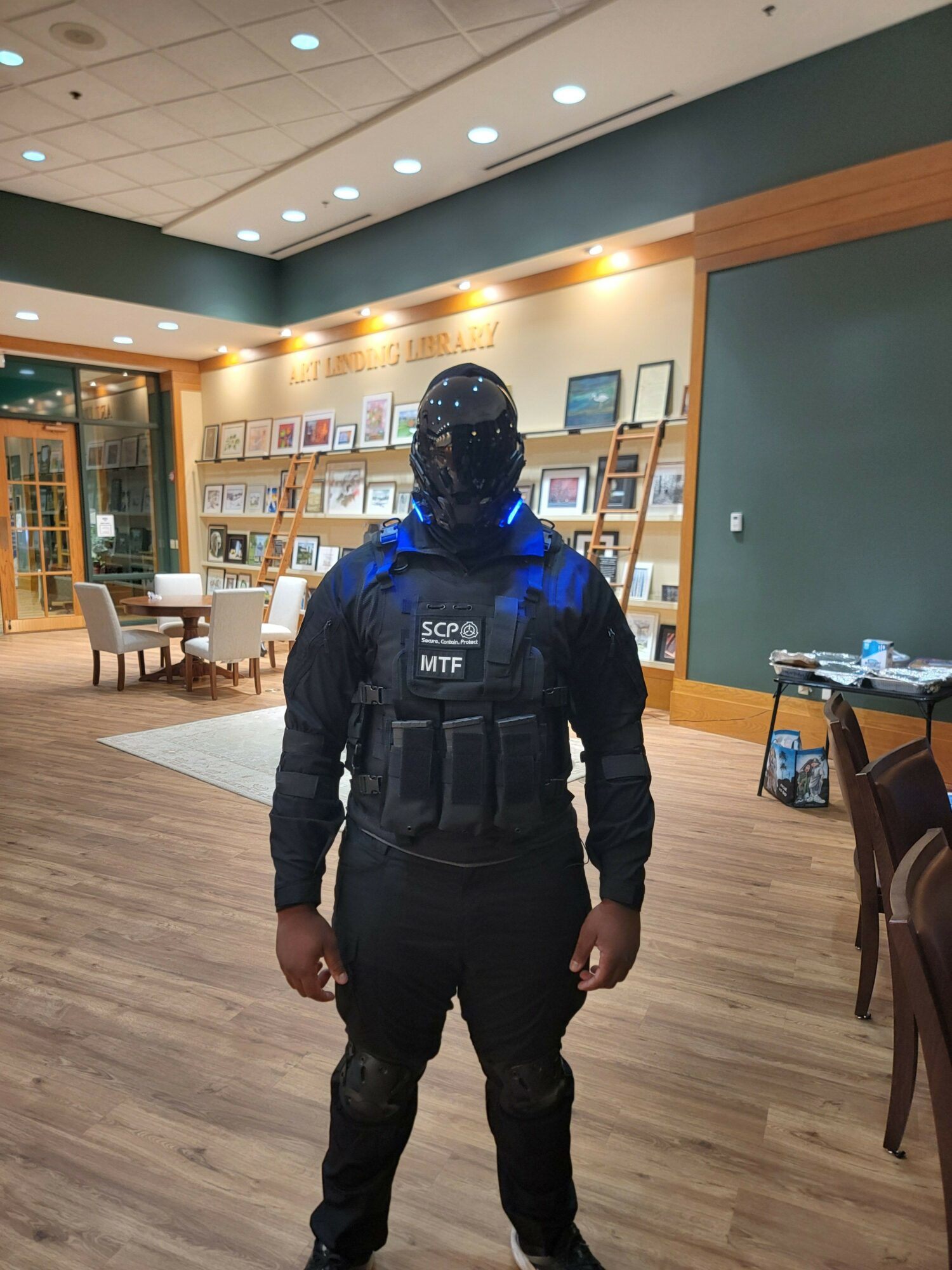 Person in tactical gear standing in a room with wooden floors and a wall of framed pictures.