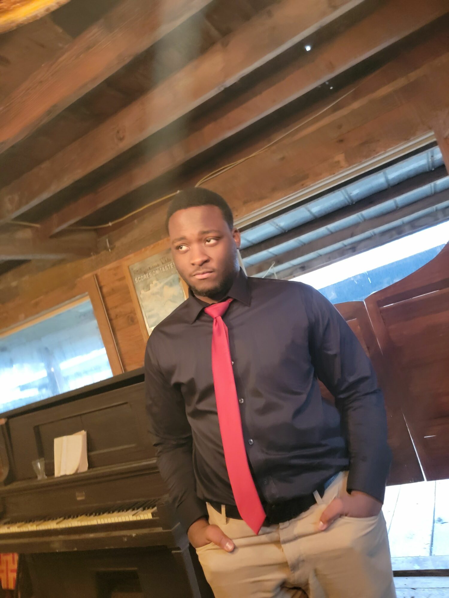 Young man in black jacket with red strap, beige pants, standing indoors near wooden beams, smiling at camera.