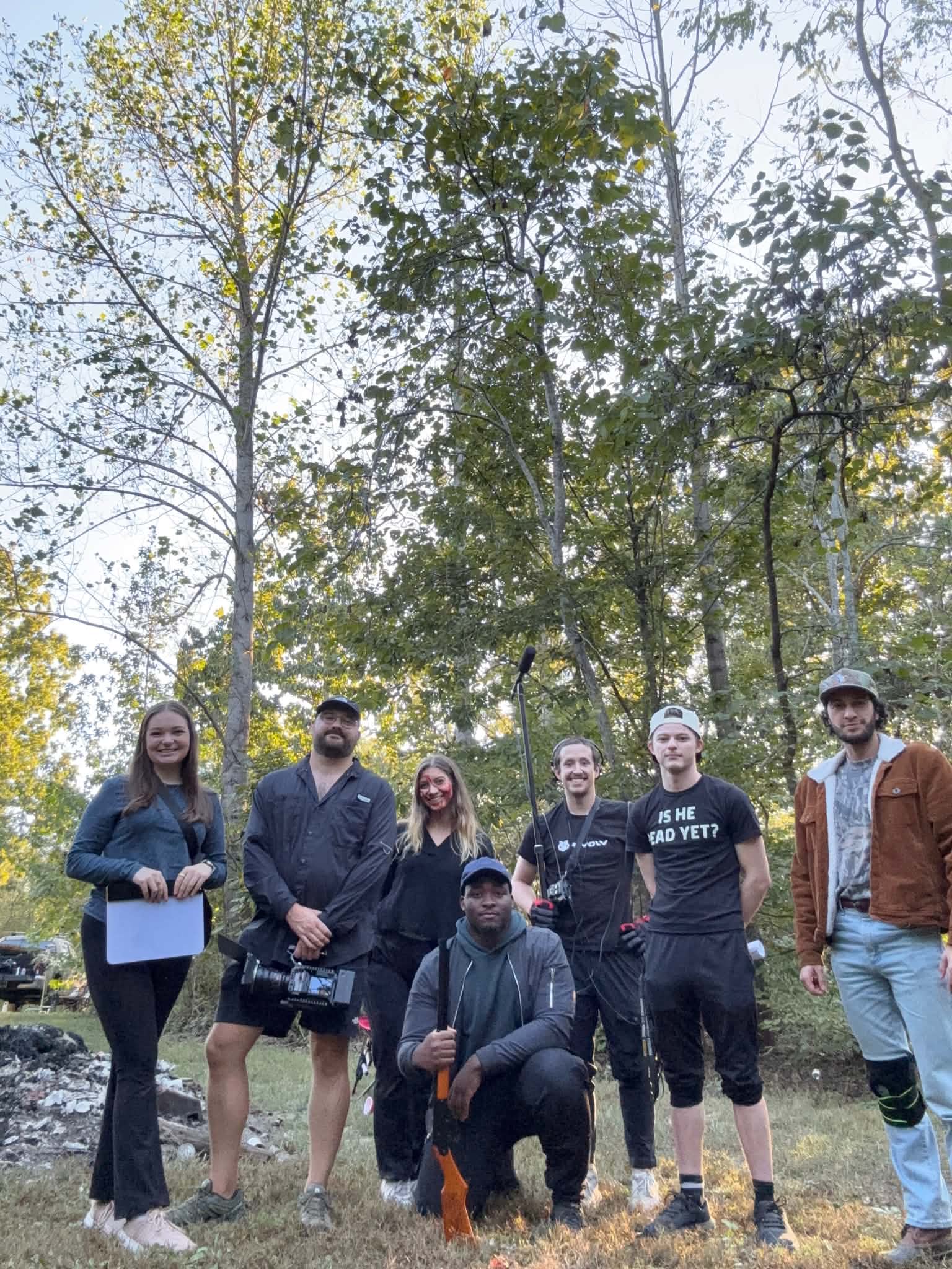 Group of seven people standing outdoors among tall trees, some holding items, smiling, in casual clothing.