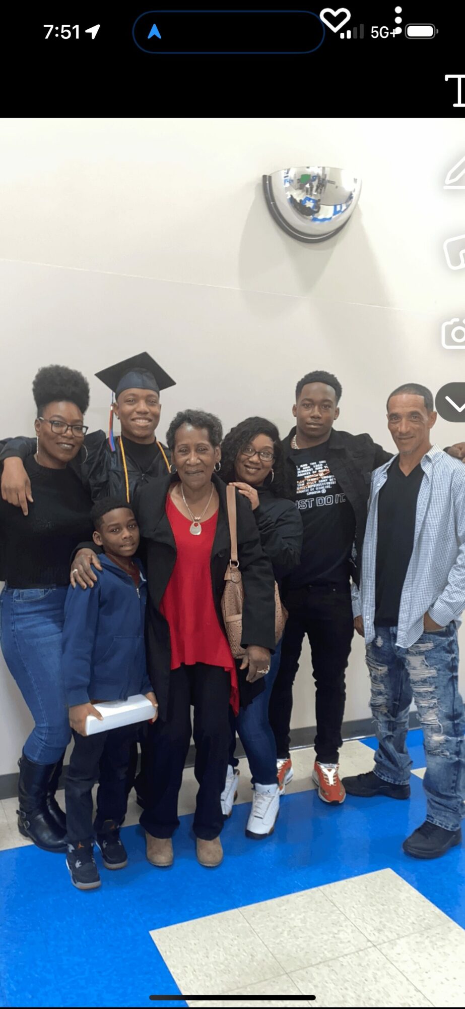 Group of six people, including a graduate in cap and gown, standing indoors against a white wall.