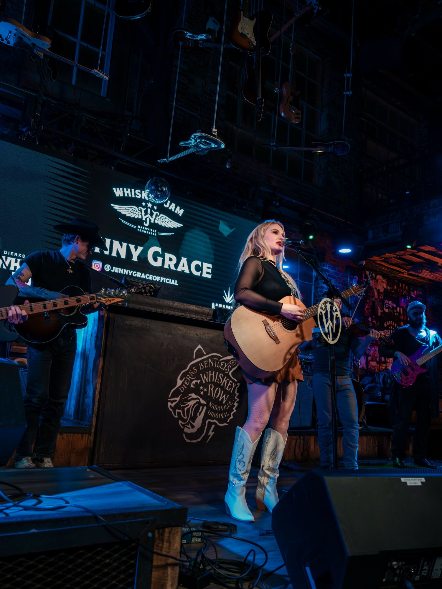 A woman with blonde hair in a black outfit and white boots plays guitar on stage with band members, a screen behind her displays text.