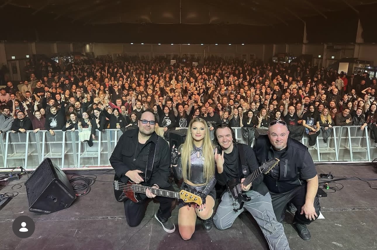 Four musicians on stage with a large audience behind them, holding guitars and posing for a photo.