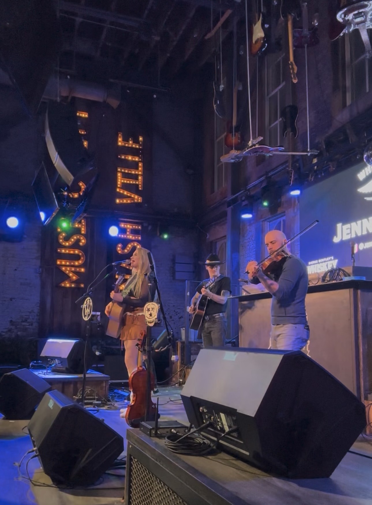Musicians perform on stage with colorful lights, a large screen, and hanging objects in the background.