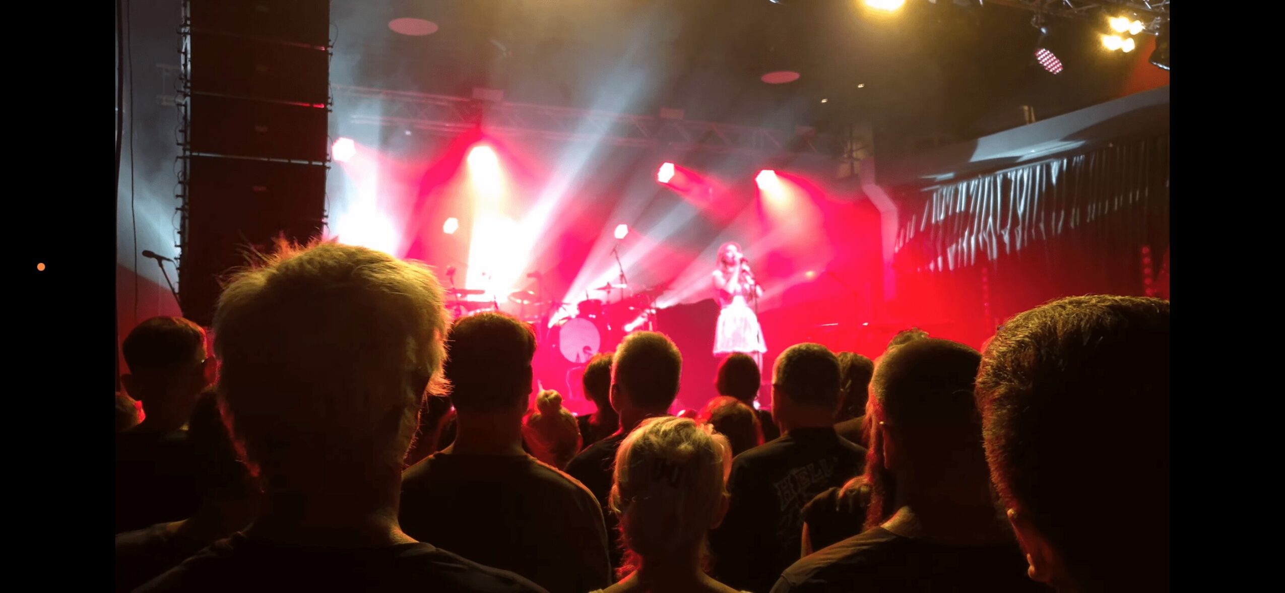 Audience watching a performer on stage with bright red and yellow lights, in a dark concert venue.