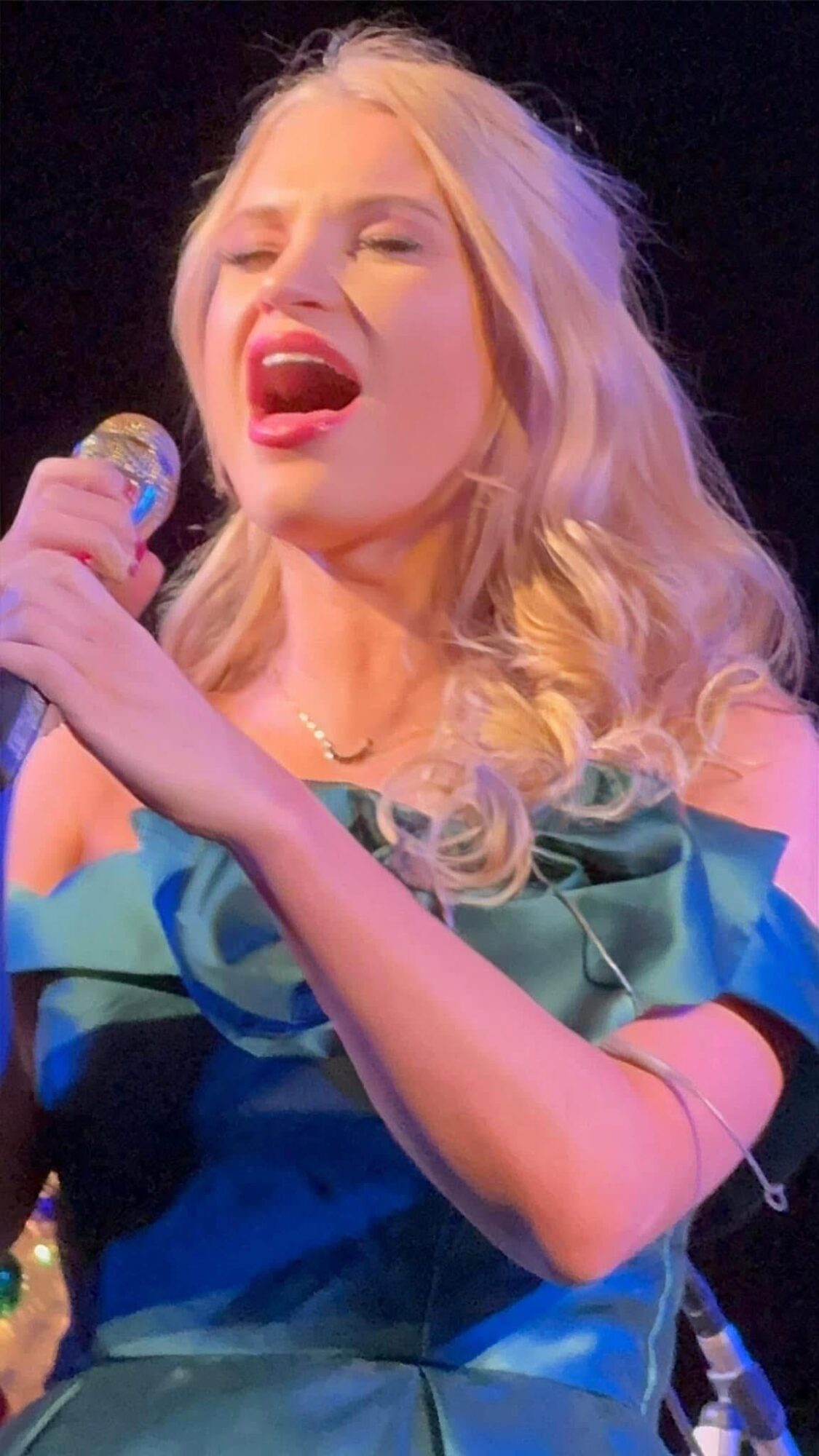 Woman singing passionately into a microphone with blonde hair and a blue dress.