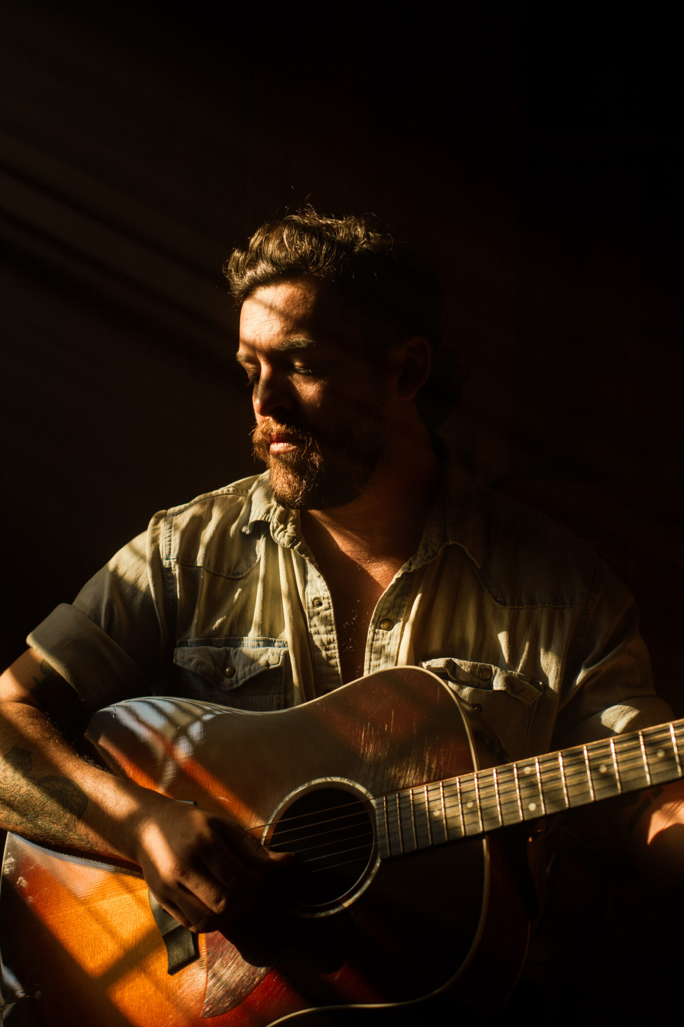 Man with beard playing an acoustic guitar in dim lighting, focused on his instrument.