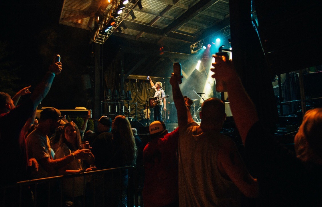 People at a concert or event with a performer on stage, some raising hands, in a dark indoor setting with stage lighting.
