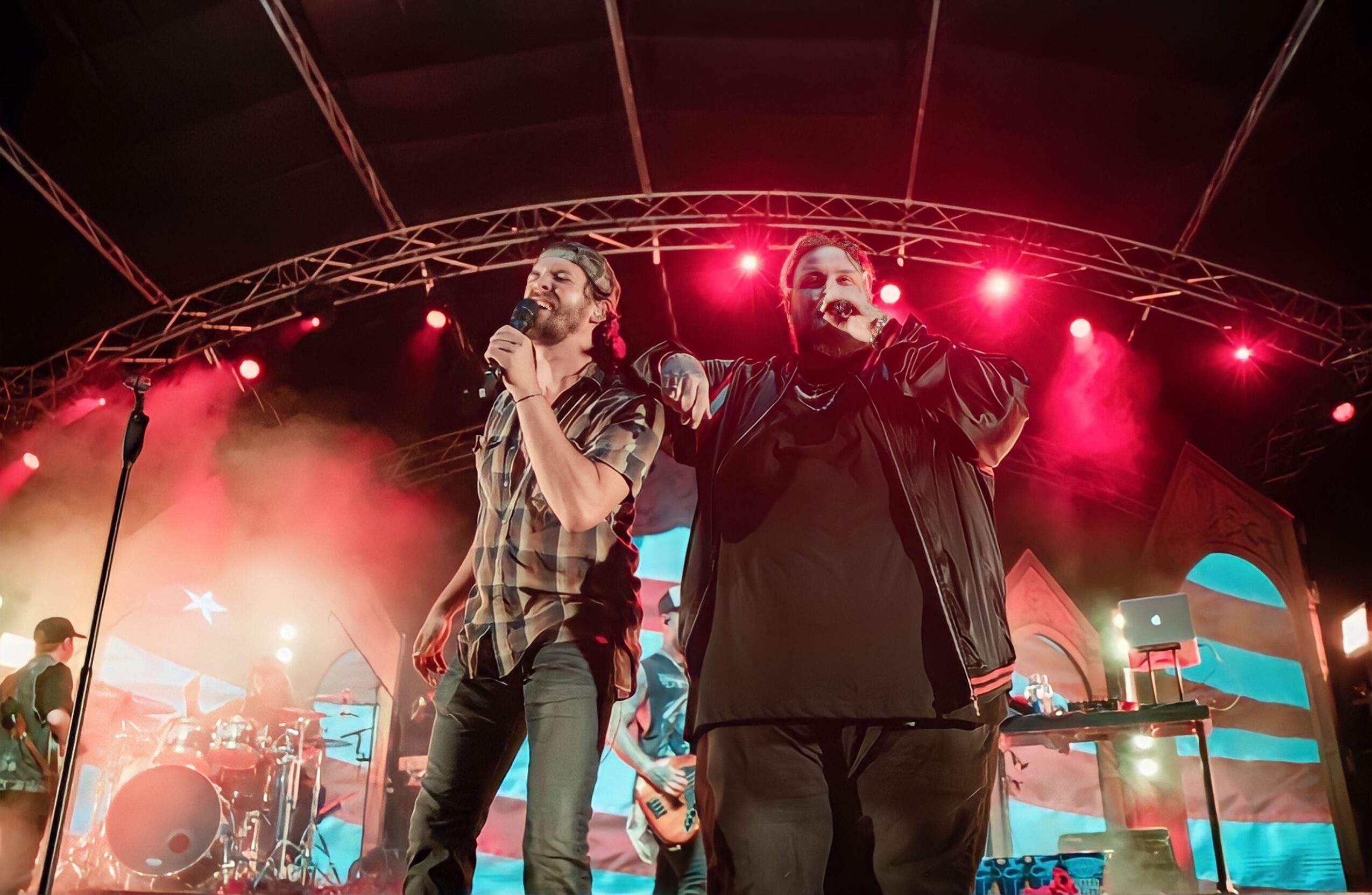 Two men sing into microphones on stage with band and red lighting in the background.