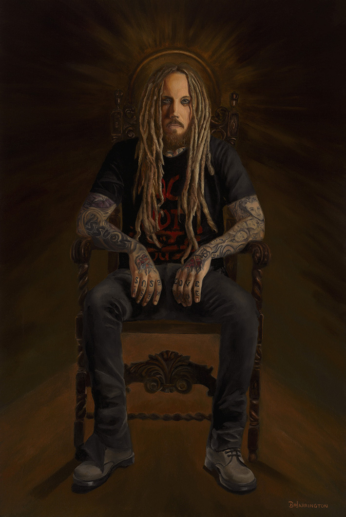 Person with long hair and tattoos sitting on a throne-like chair, dark background, serious expression.