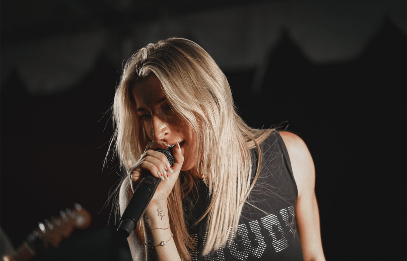 A woman with long blonde hair singing into a microphone on stage, wearing a sleeveless black top.
