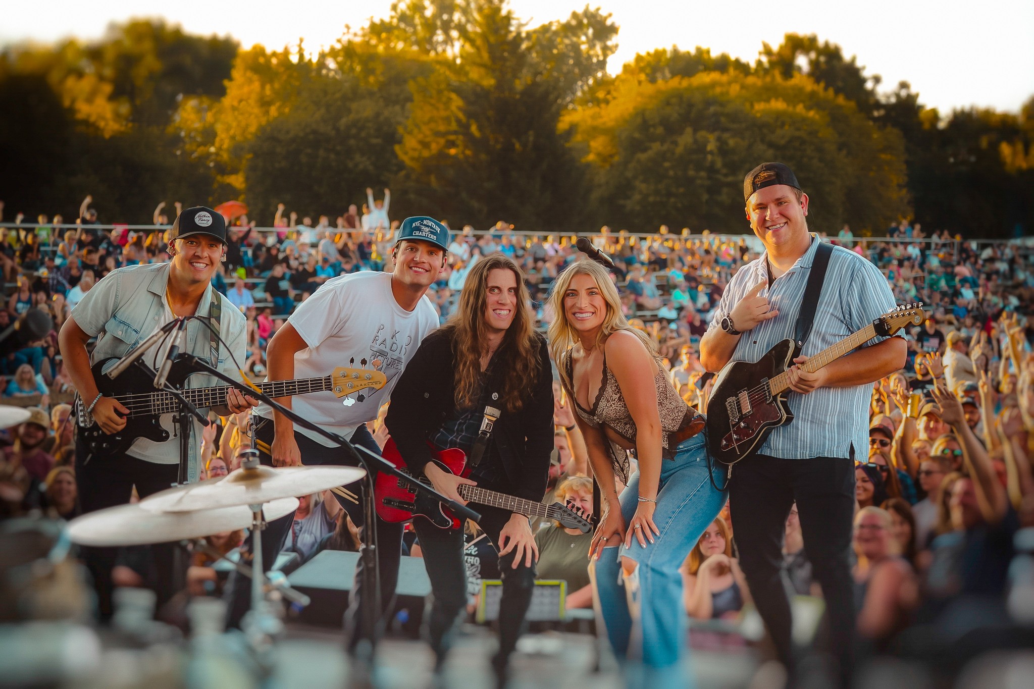 Five musicians perform on stage with a large crowd and trees in the background, during an outdoor concert.