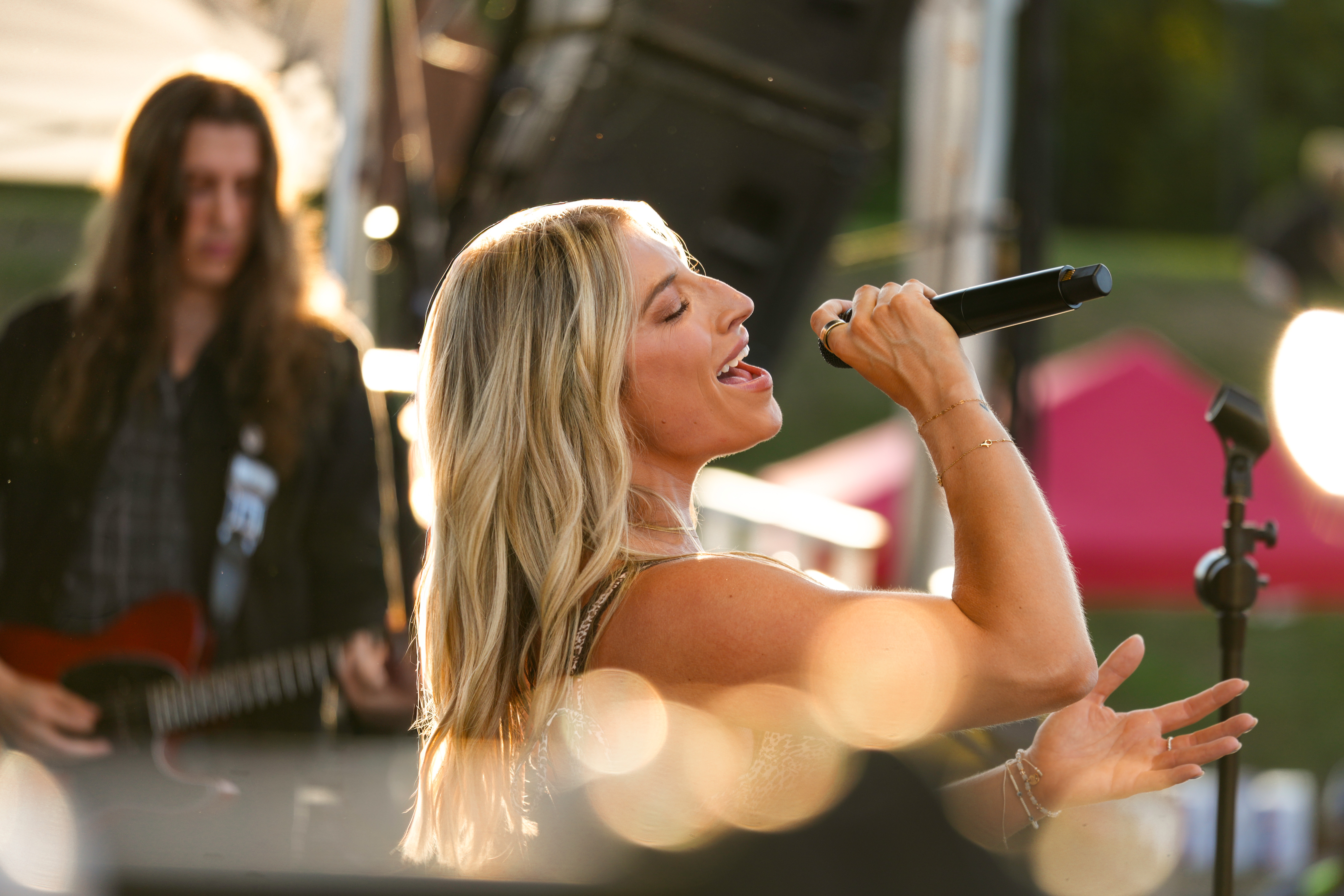 Woman singing into a microphone outdoors with a guitarist in the background, sunlight and blurred background.