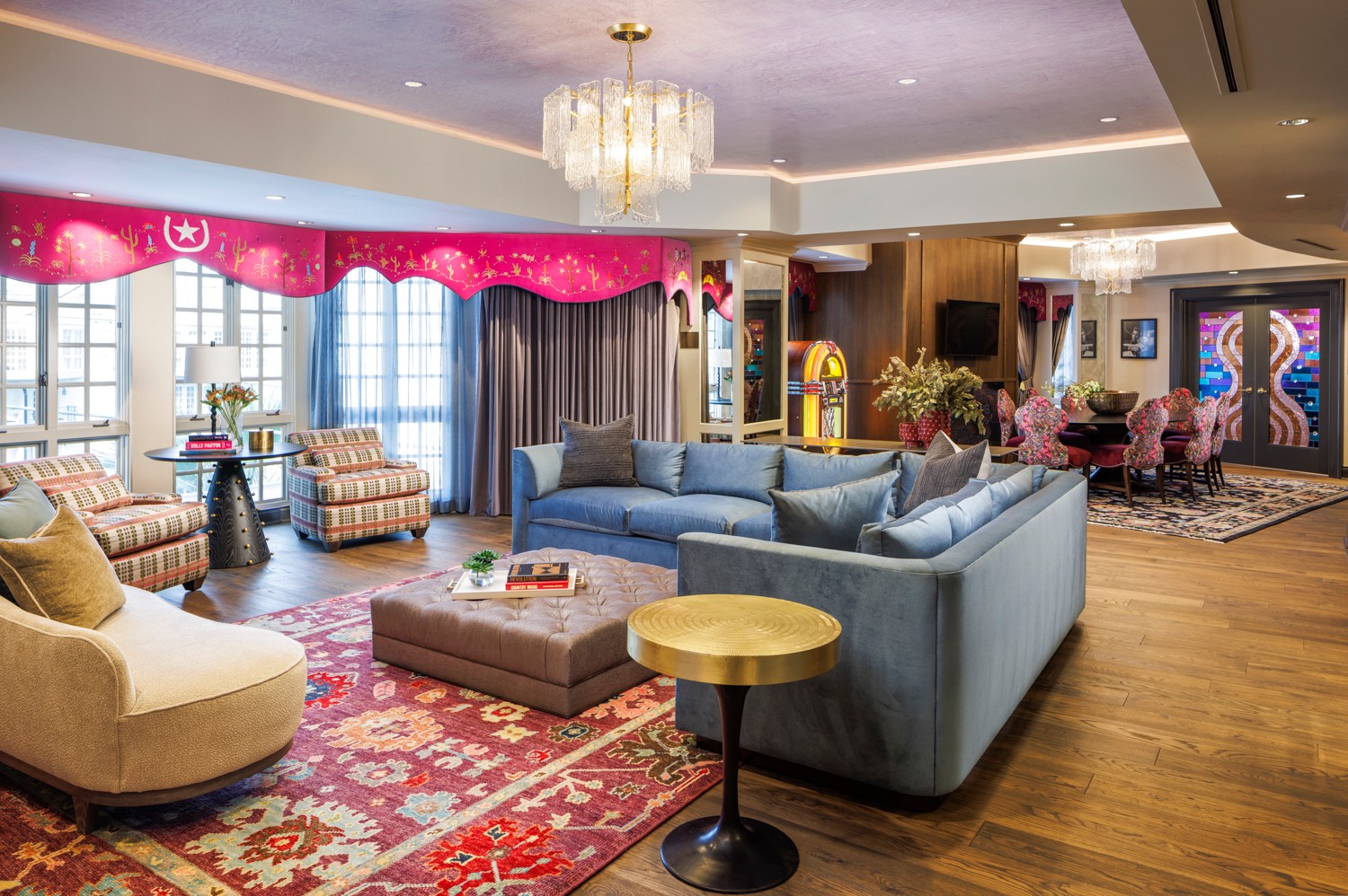 Living room with blue and beige sofas, a red patterned rug, large windows, and pink curtains, decorated with flowers and artwork.
