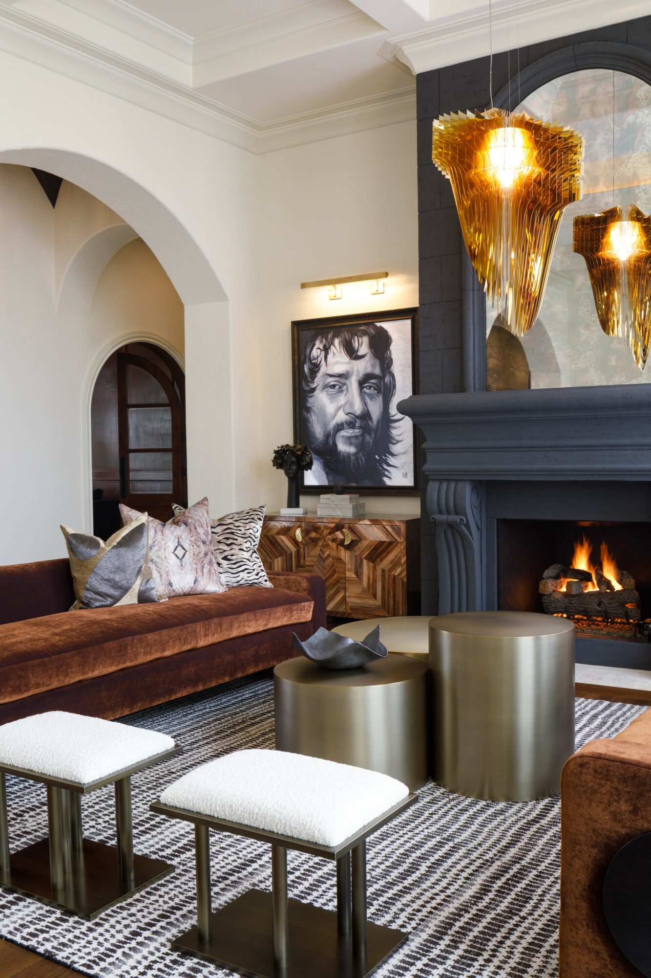 Living room with a fireplace, portrait of a man, gold pendant lights, and seating area with stools and cushions.