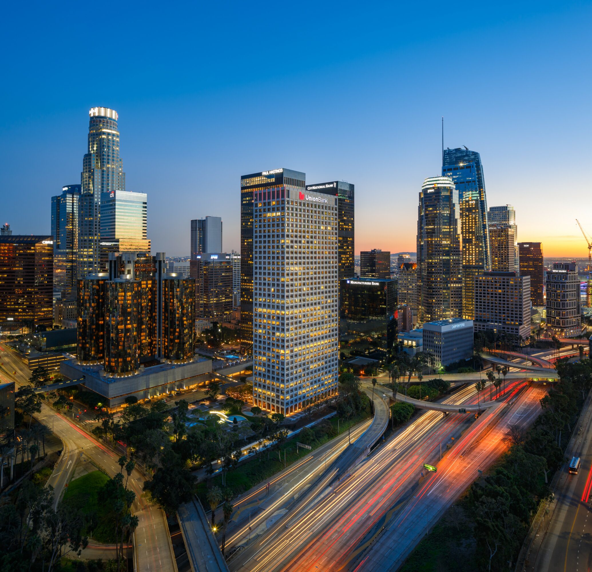 City skyline with tall buildings during dusk, light trails on roads, and a clear sky.