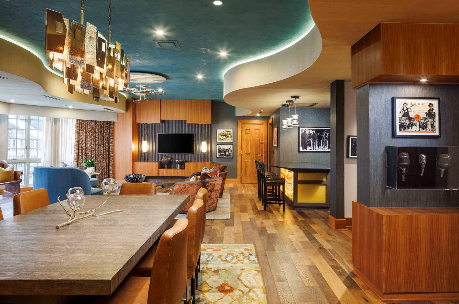 Interior of a modern lounge with a large wooden table, chairs, a TV, artwork, and a colorful ceiling design.
