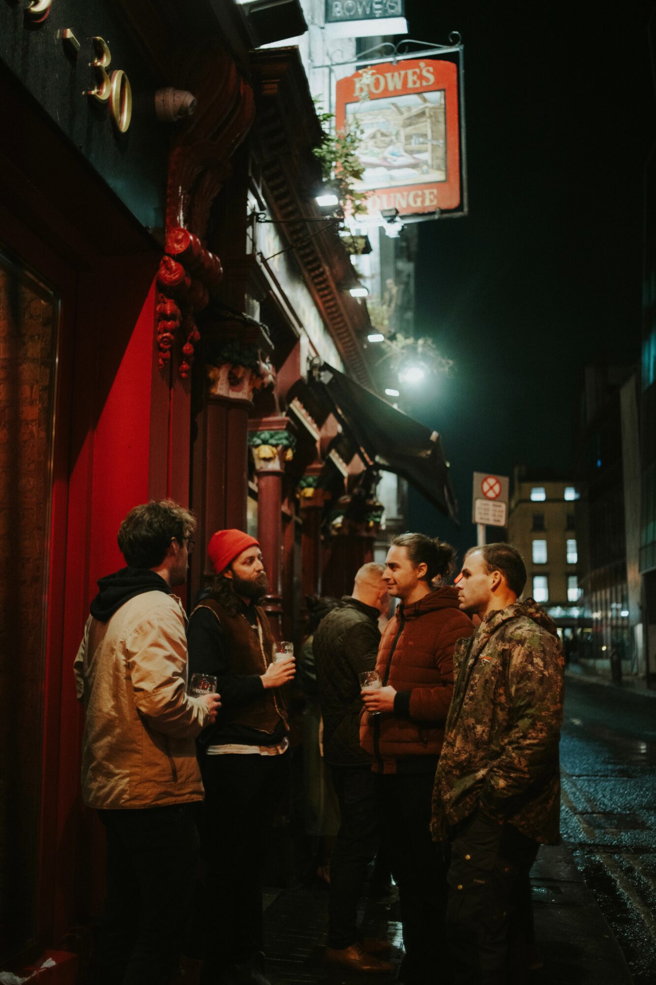 Group of five people standing outside a building at night, talking, with streetlights and signs visible in the background.
