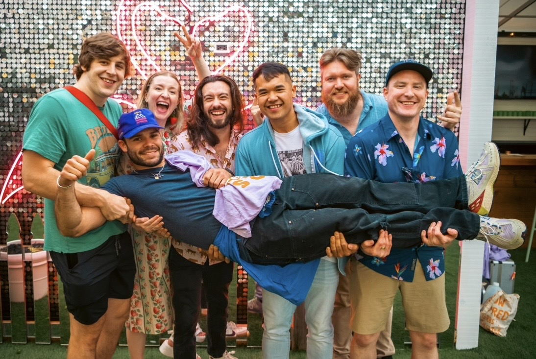 Group of six people holding a person horizontally, smiling, in front of a colorful background with lights and decorations.