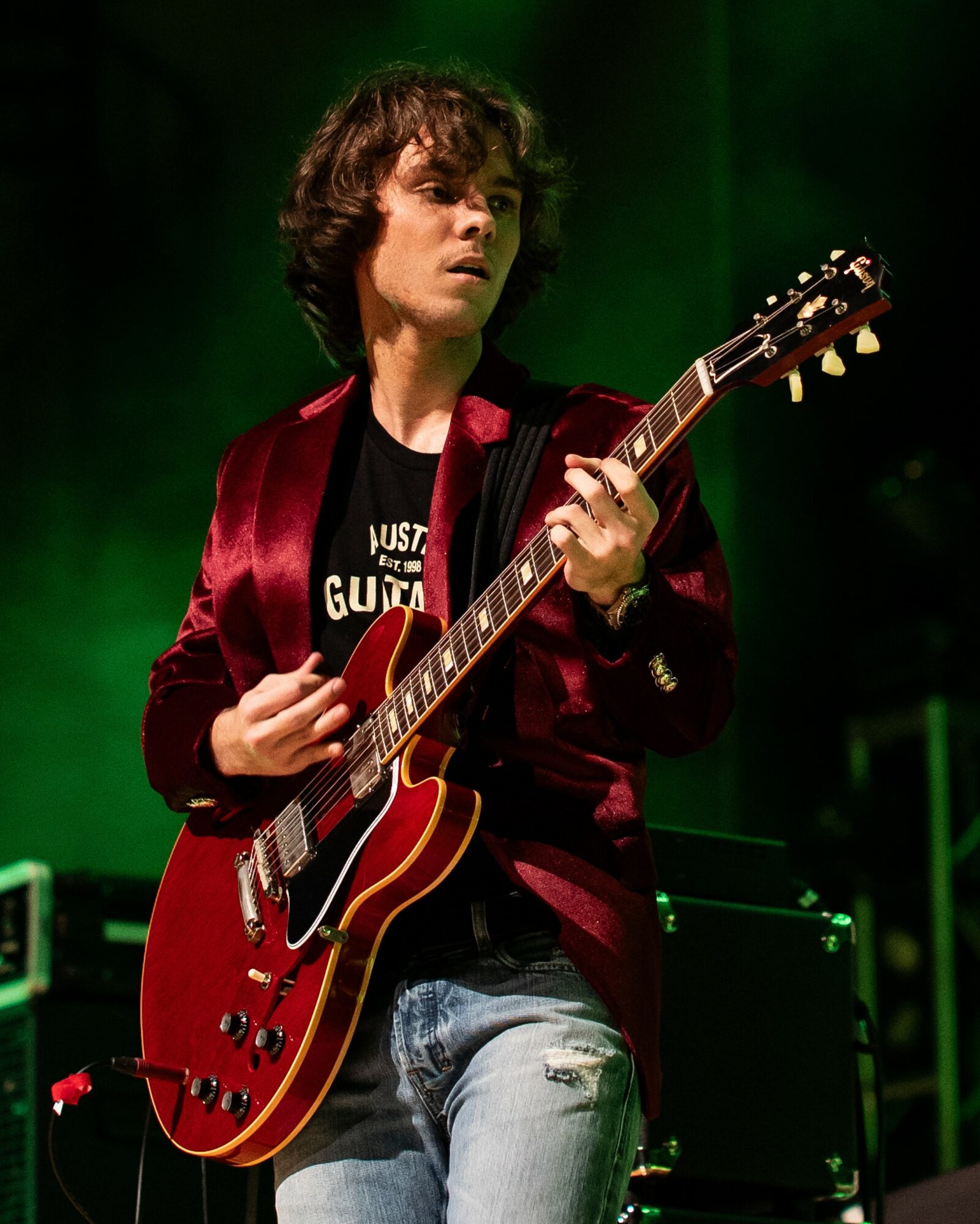 Person with curly hair playing a red electric guitar on stage with green lighting, wearing a dark shirt and red jacket.
