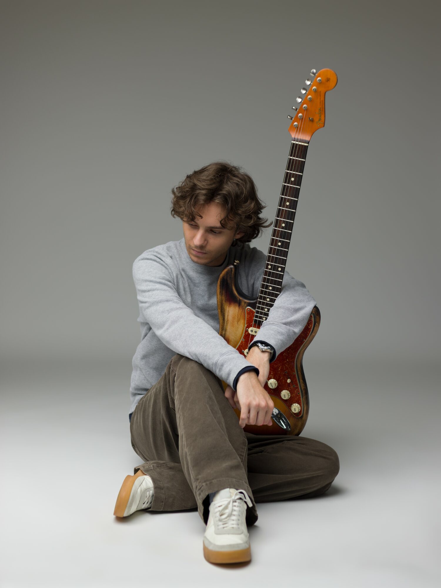 Person sitting on the floor holding an electric guitar, looking down, with a plain gray background.