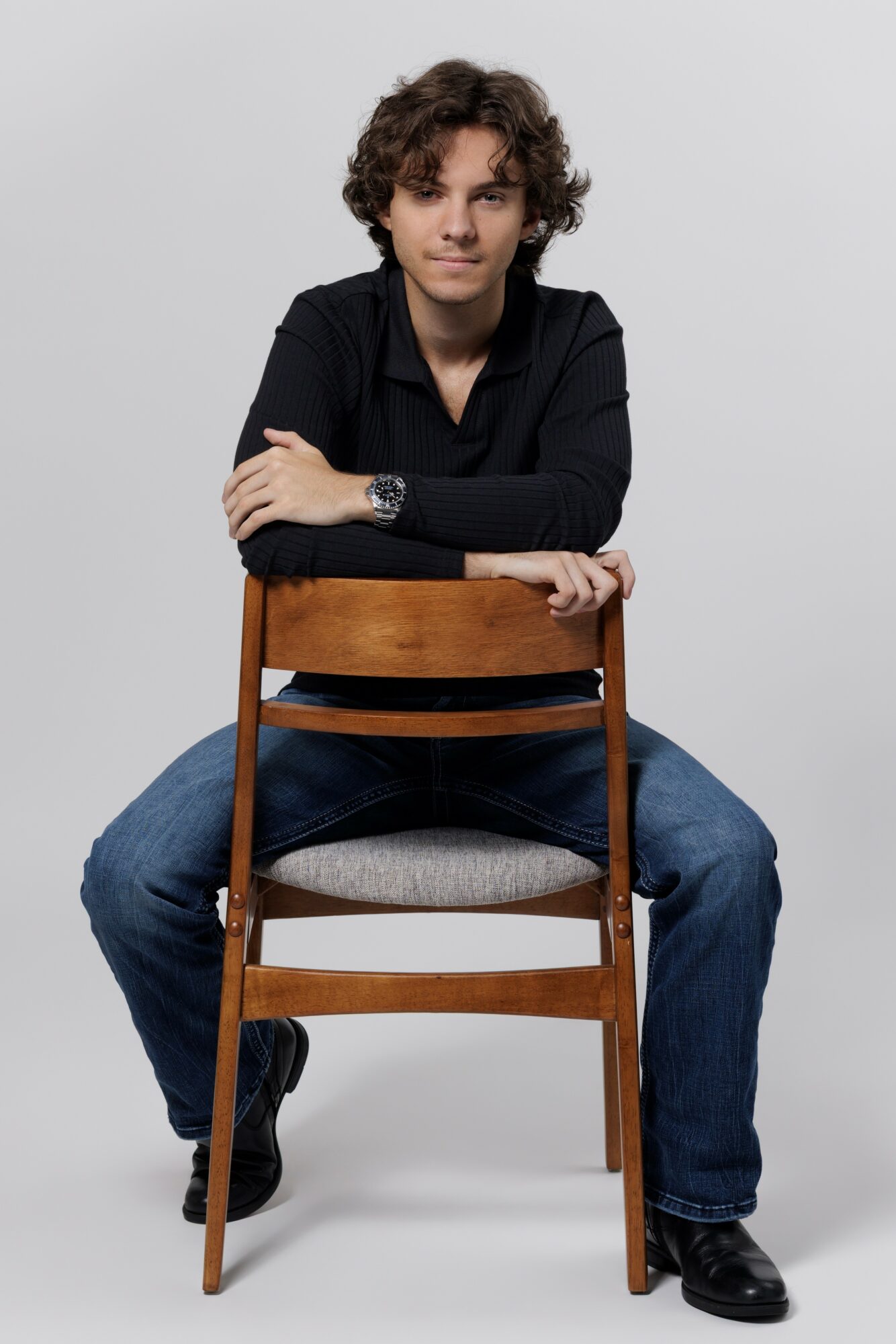 Person with curly hair sitting on a chair, arms crossed, wearing a black shirt and jeans, against a plain background.