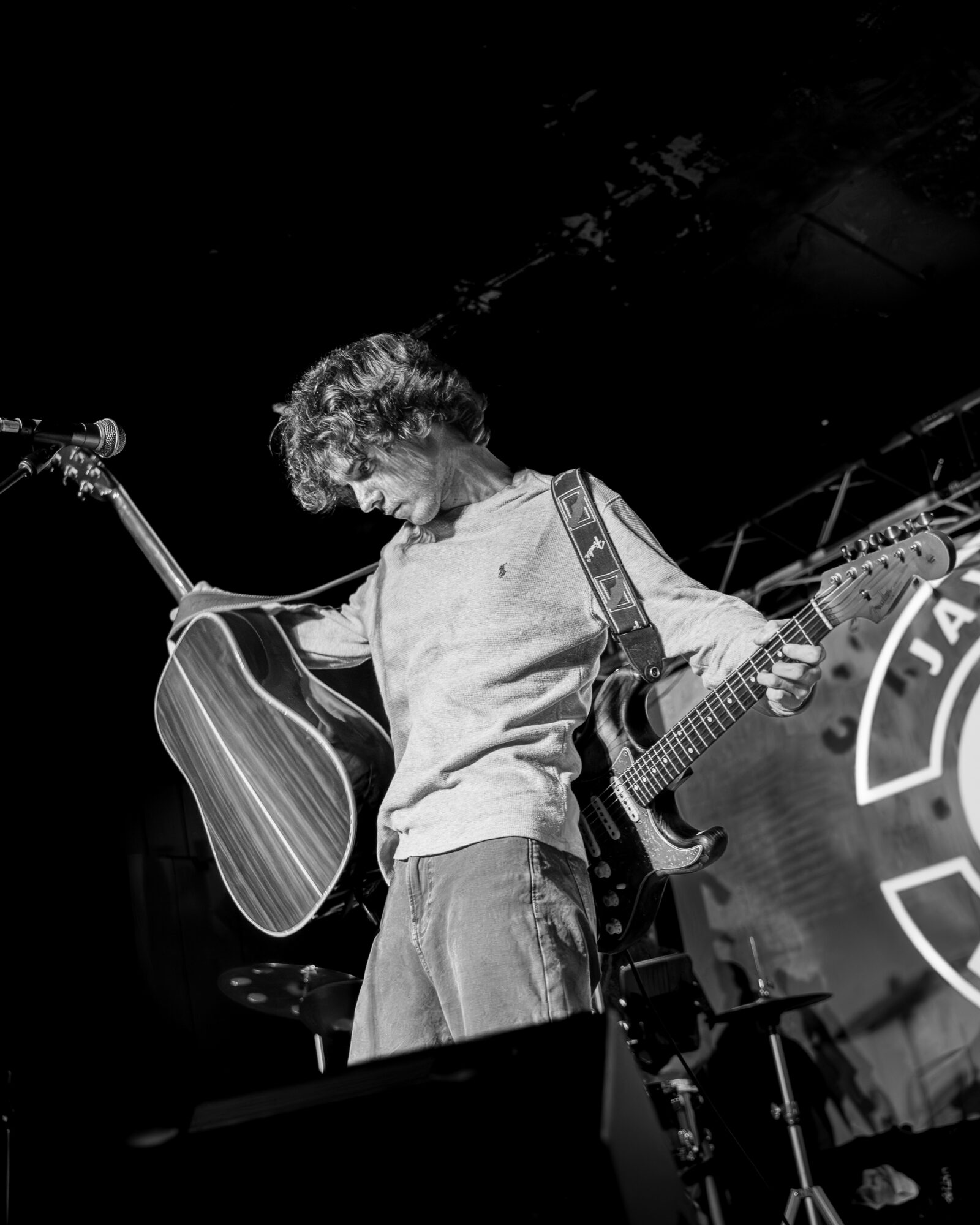 Person with curly hair holding an acoustic guitar on stage, with a drum set and a logo in the background.